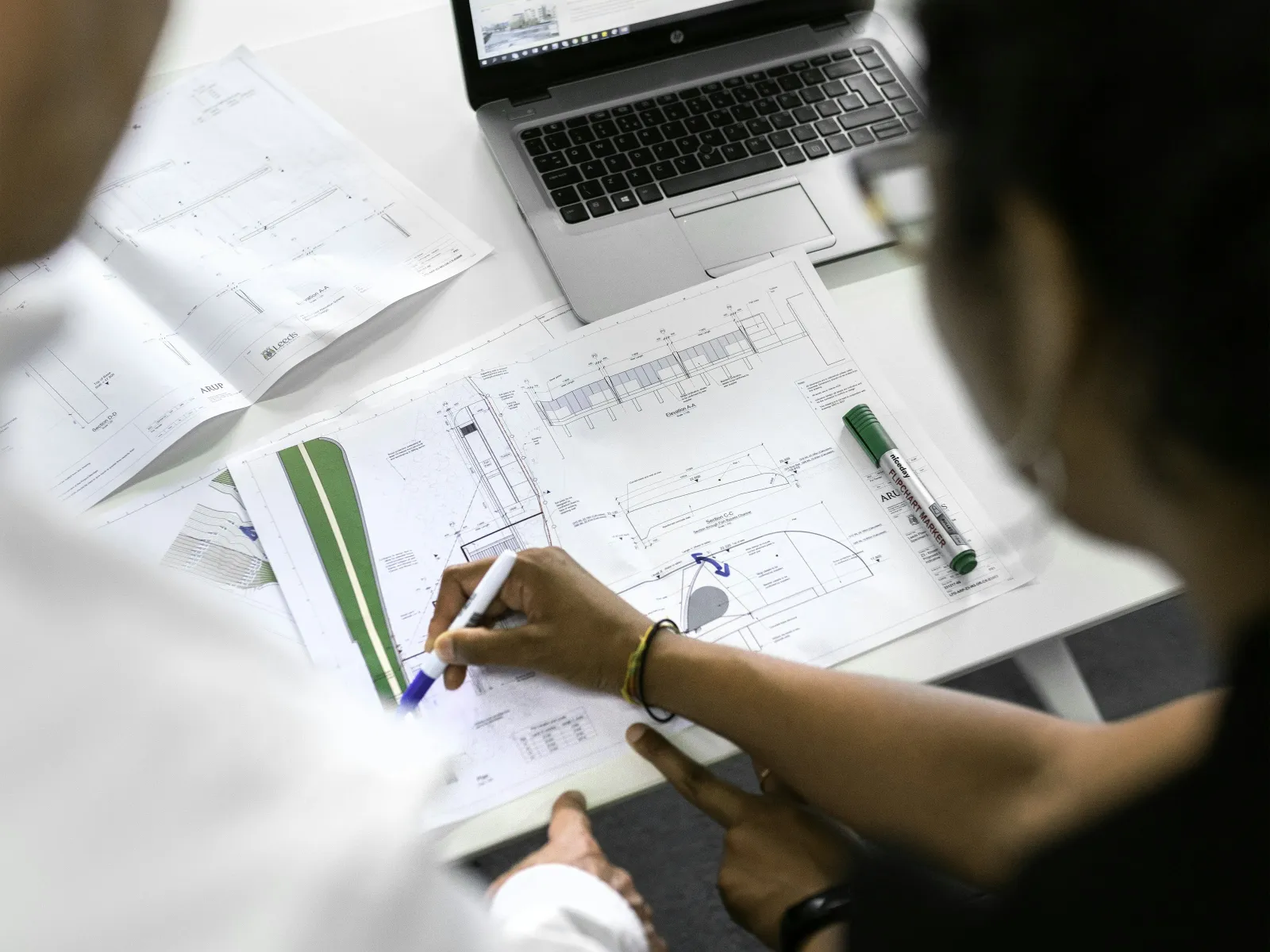 Two professionals reviewing architectural plans and designs on paper and laptop during a work meeting.