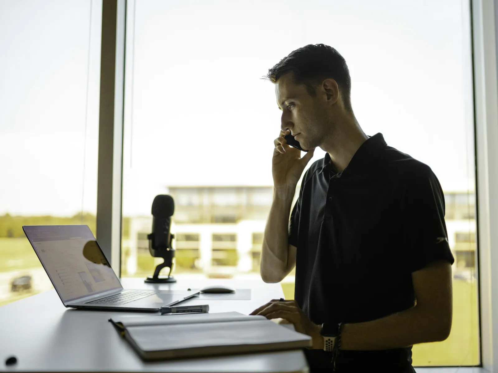 Man in blue shirt thoughtfully working on laptop in bright modern office space with blurred windows.