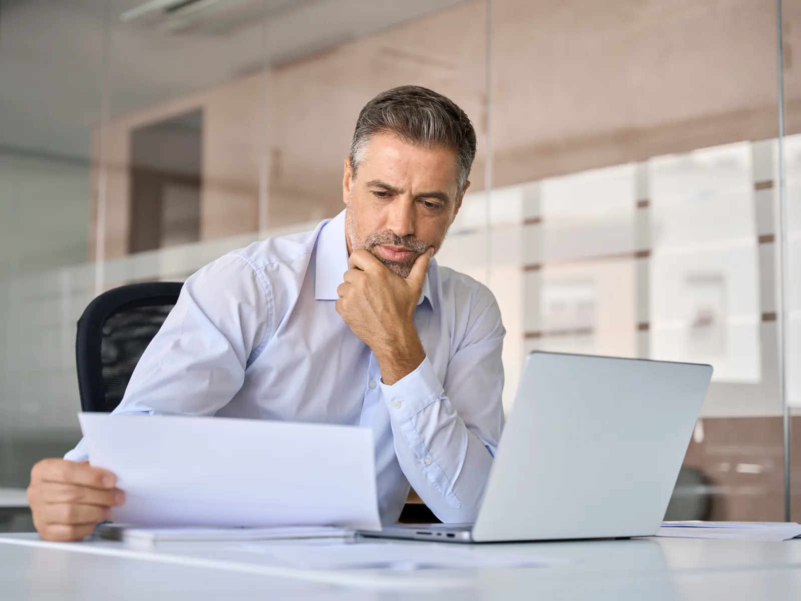 Businessman in white shirt thoughtfully reviewing documents at desk with laptop in modern office