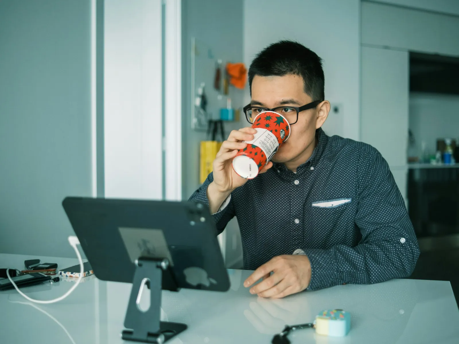 Man wearing glasses drinking from a decorative cup while using a tablet on a white desk in a modern kitchen.