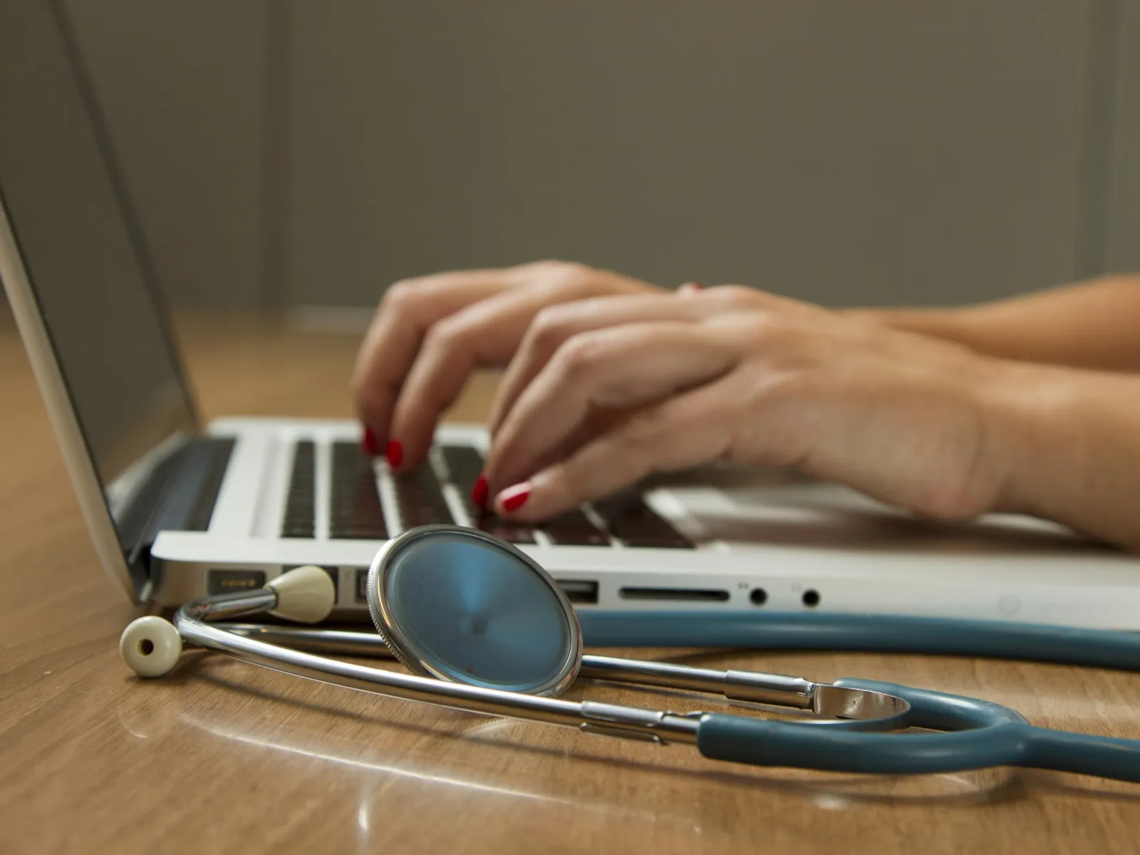 Close-up of hands typing on a laptop with a stethoscope on a wooden desk symbolizing medical work.