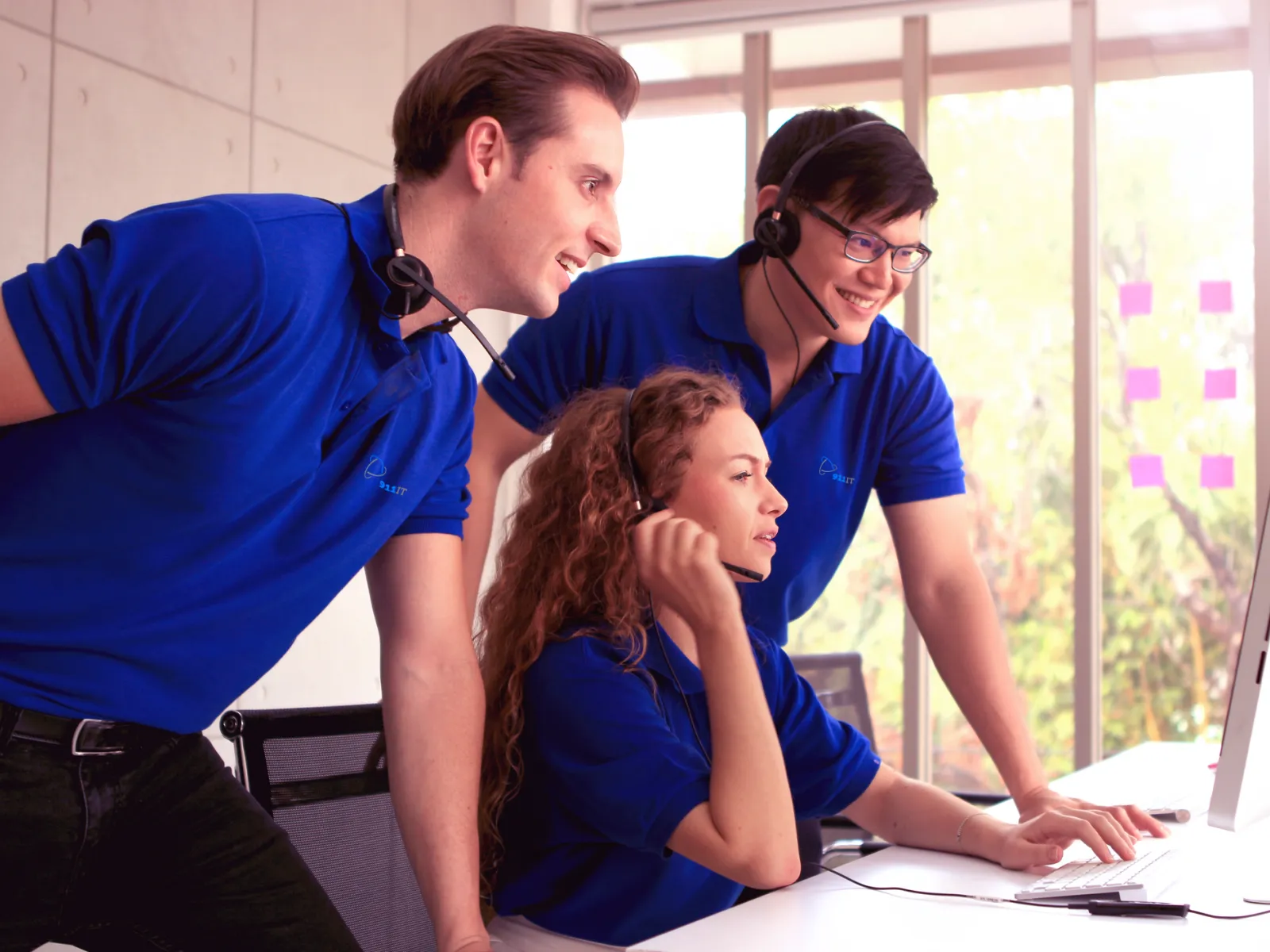 Three customer service representatives in blue polos collaborate while looking at a computer in a bright office.
