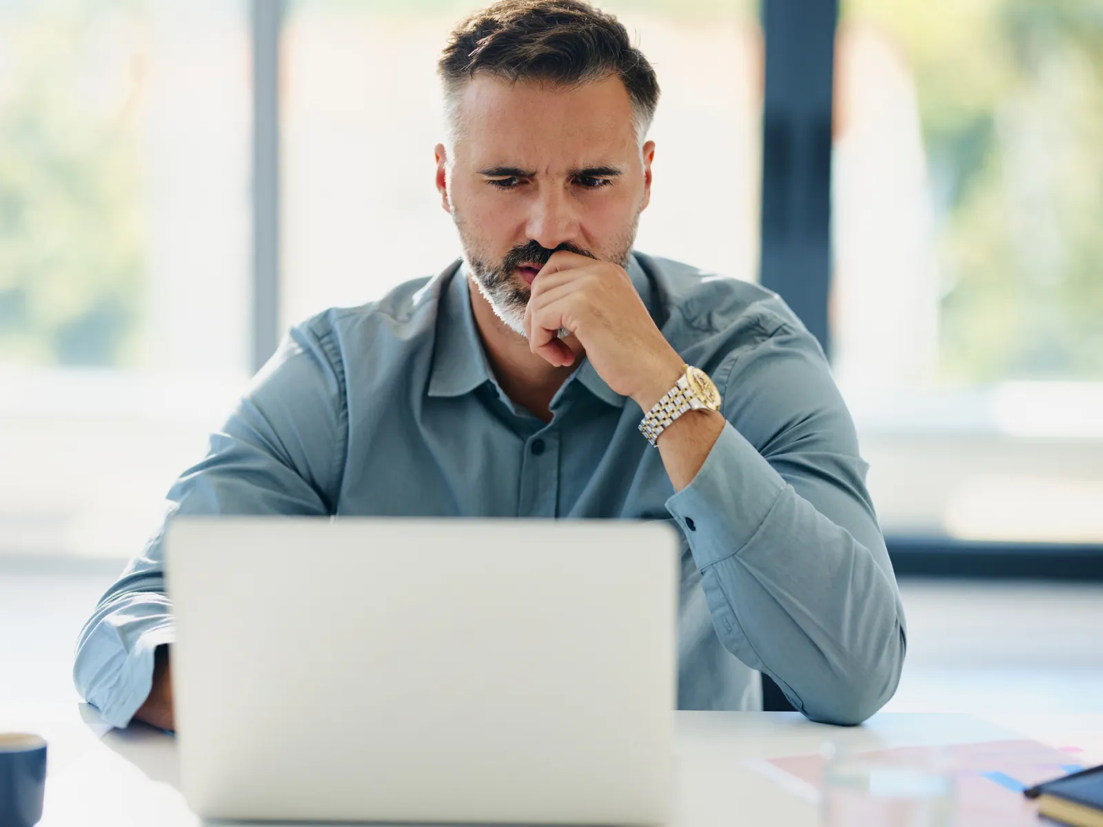 Man in blue shirt thoughtfully working on laptop in bright modern office space with blurred windows.
