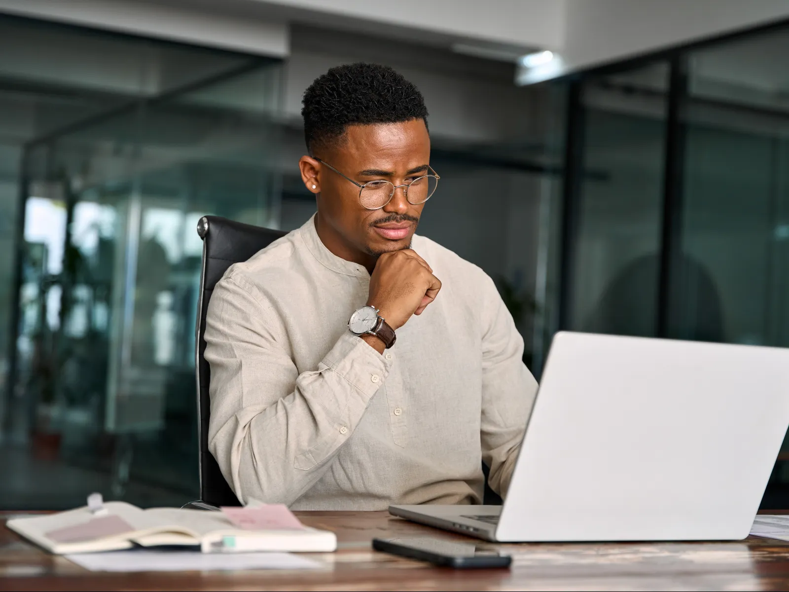 Man wearing glasses focused on laptop screen working at office desk with notebook and smartphone nearby