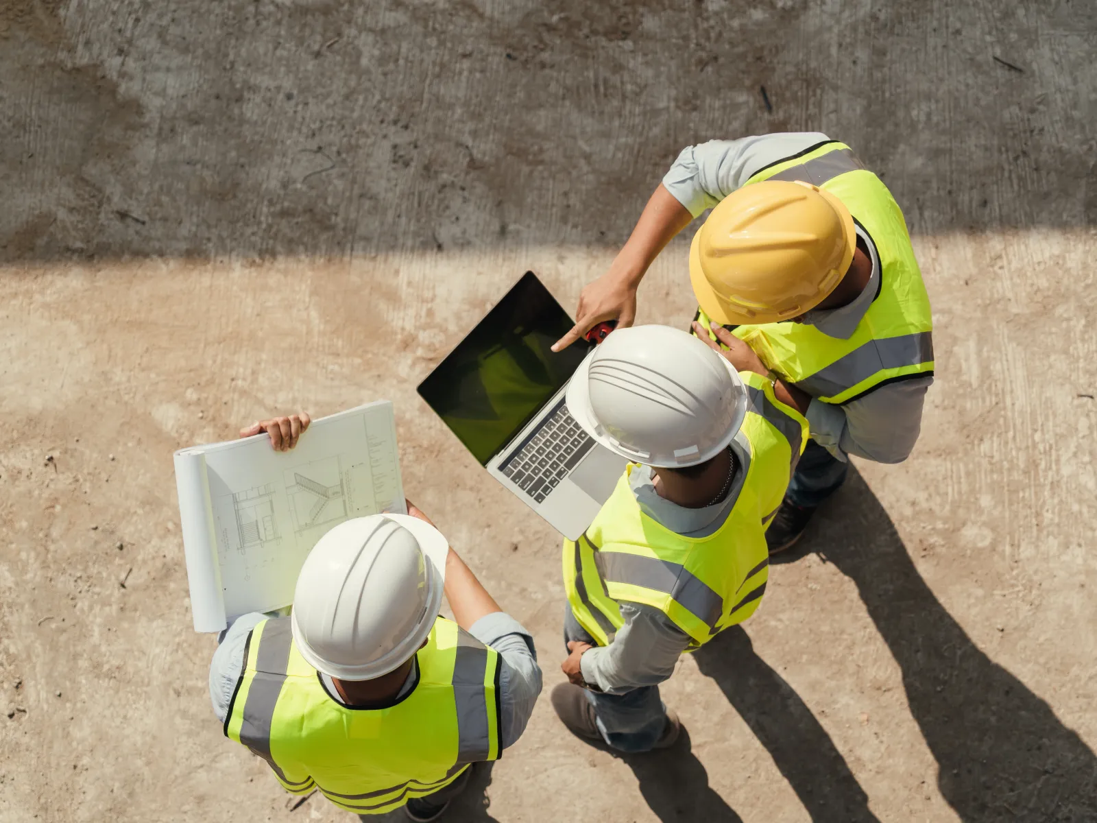 Overhead view of three construction workers wearing safety helmets and vests reviewing plans and a laptop on site.