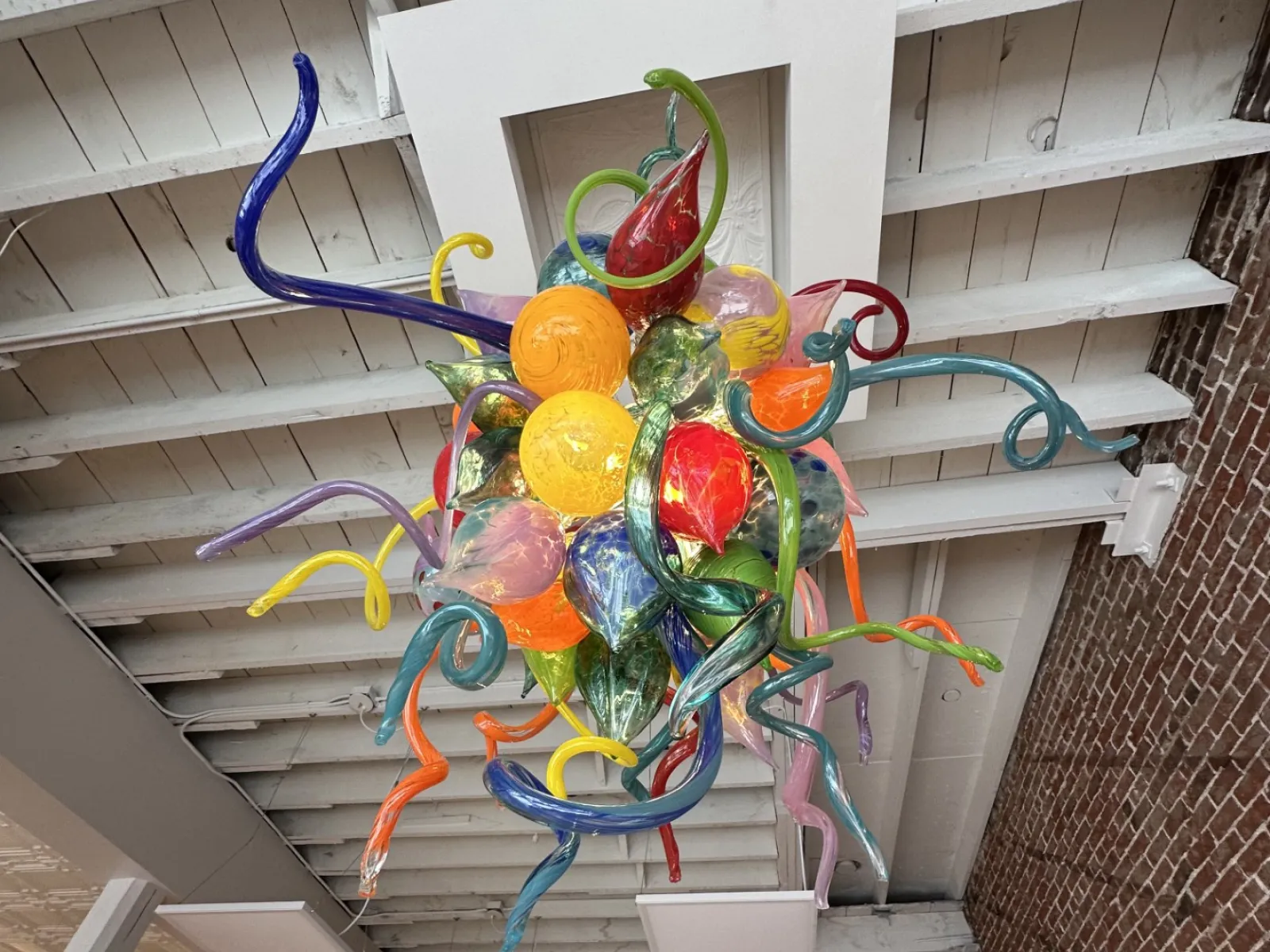 Colorful glass chandelier with twisted, organic shapes hanging from a white wooden ceiling in an art gallery.