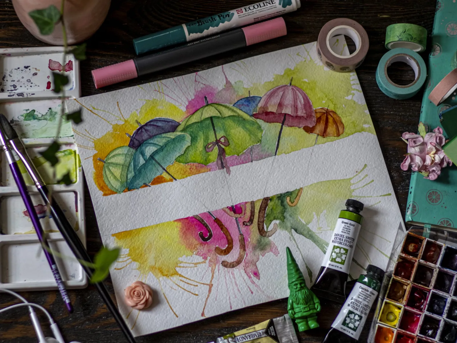 Watercolor painting of colorful umbrellas surrounded by painting supplies and brushes on wooden table.