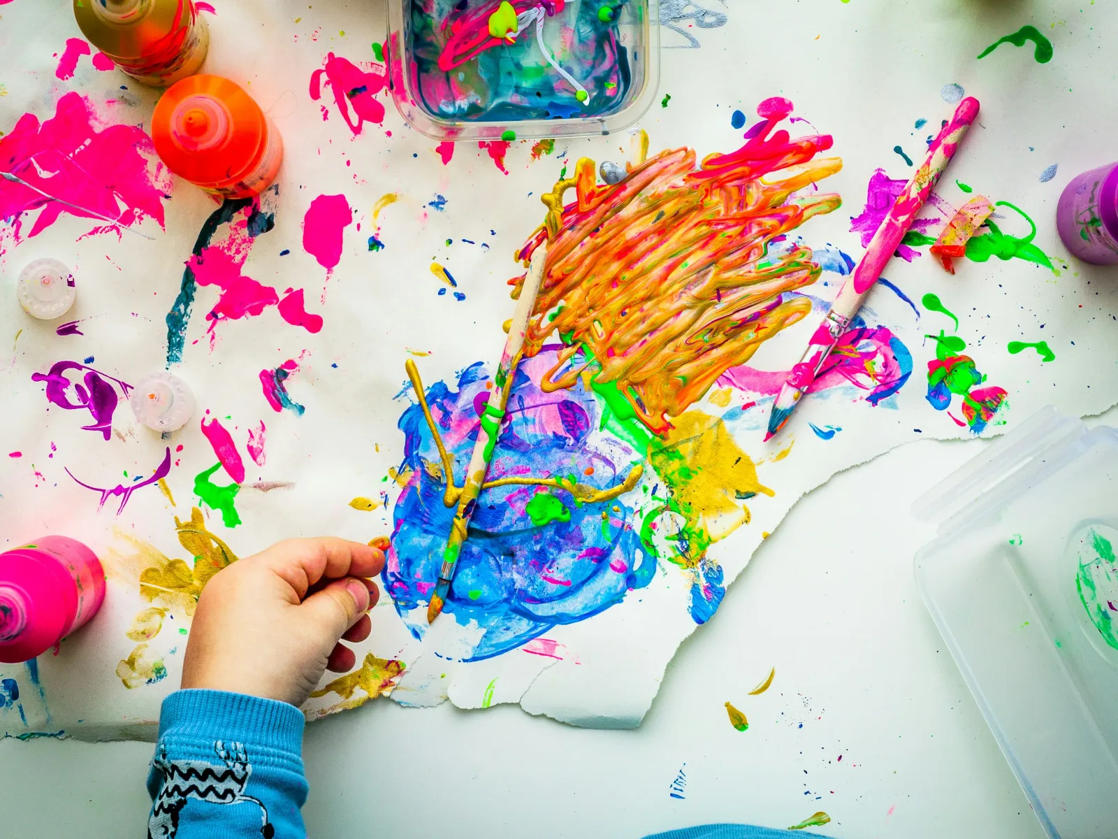 Child's hand painting colorful abstract art with bright paints and sticks on white paper.