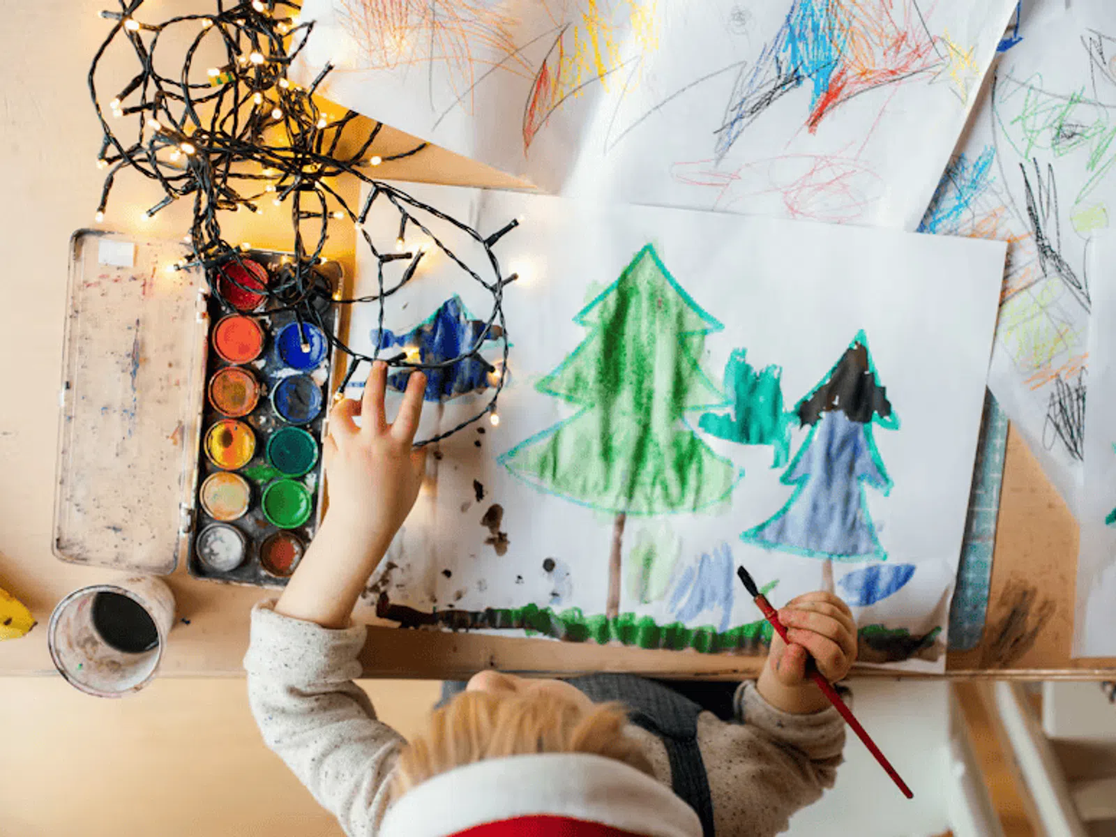 Child painting colorful trees with watercolors at a table decorated with fairy lights and drawings.