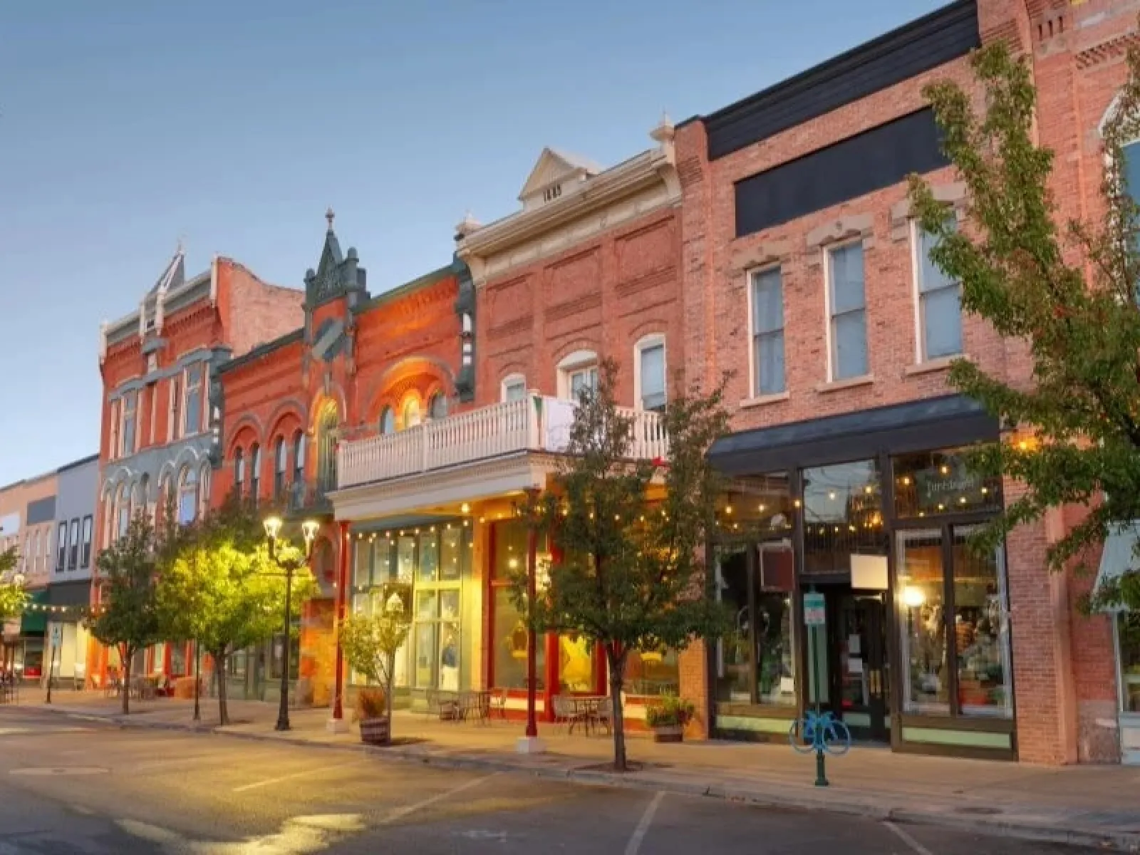 Historic downtown street with brick buildings, trees, street lamps, and storefronts at dusk in warm lighting