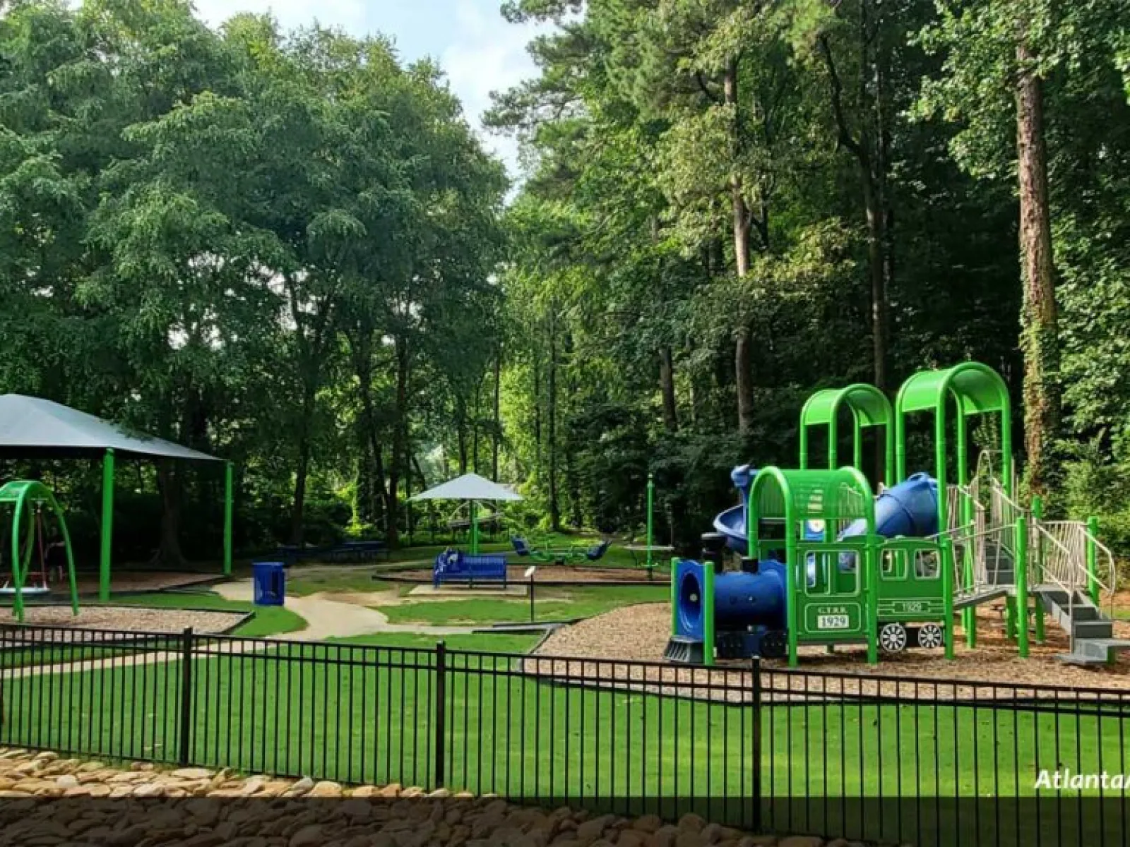 Children's playground with green and blue train-themed equipment, swings, benches, and shaded areas surrounded by trees.