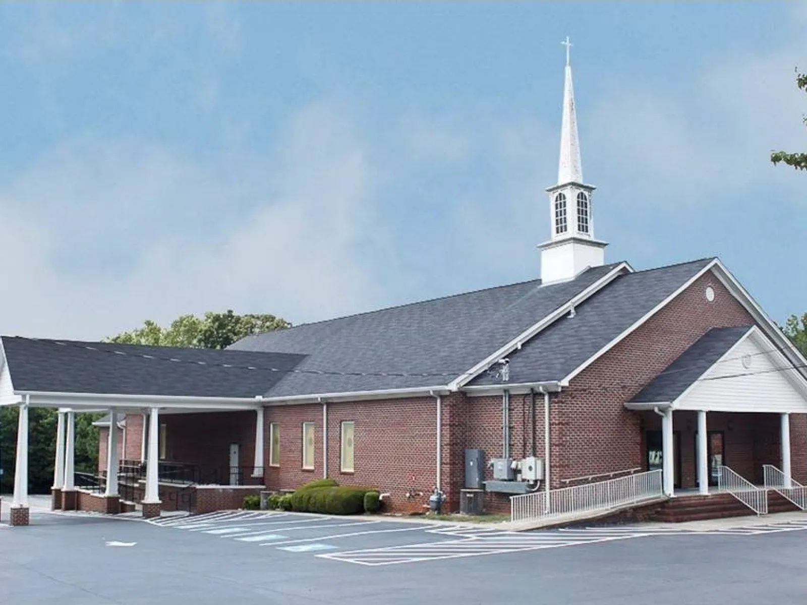Brick church building with white steeple, covered entrance, and wheelchair ramps under a clear sky.