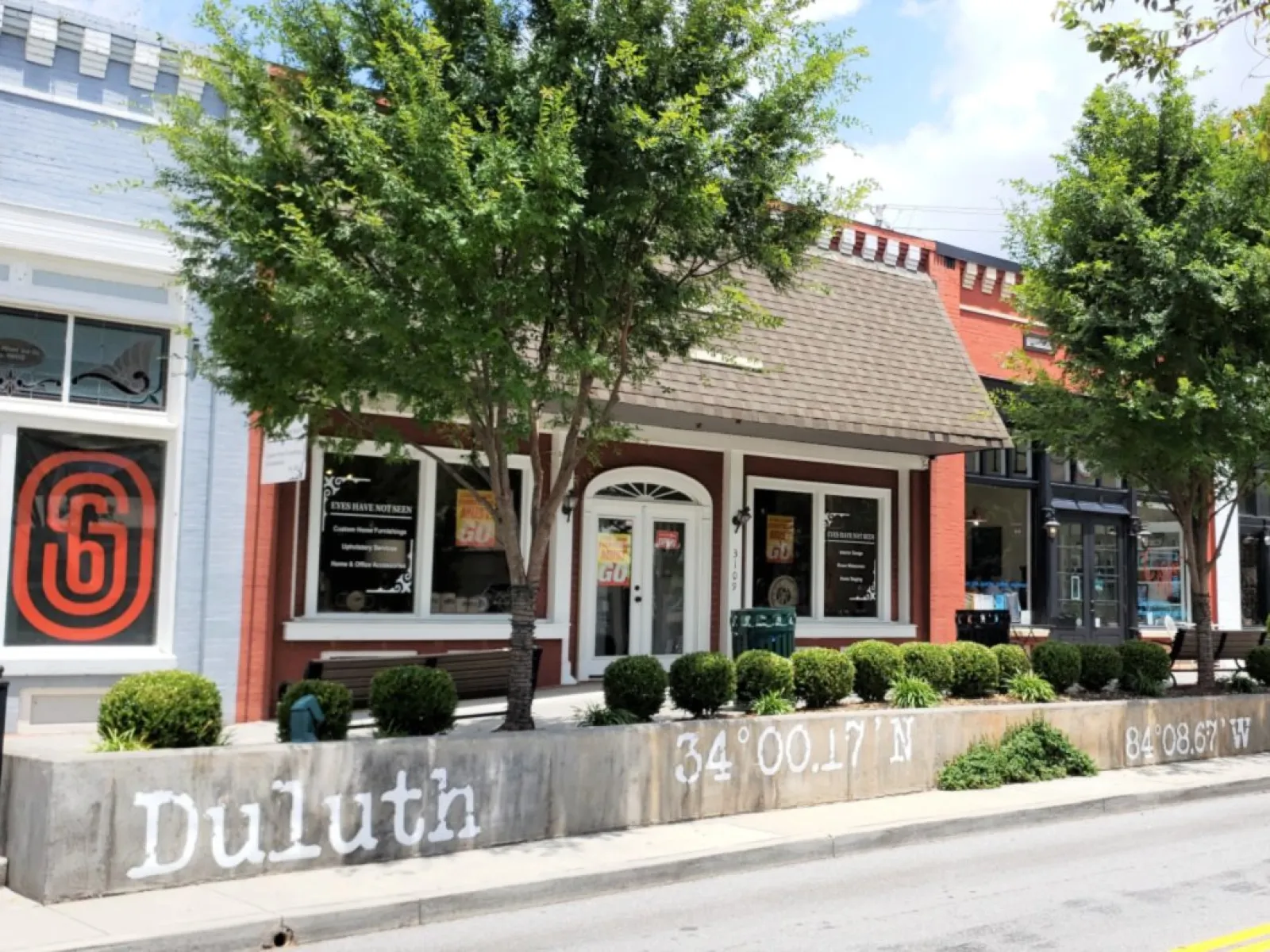 Duluth street view showing storefronts, trees, and coordinates painted on a concrete barrier under a sunny sky.