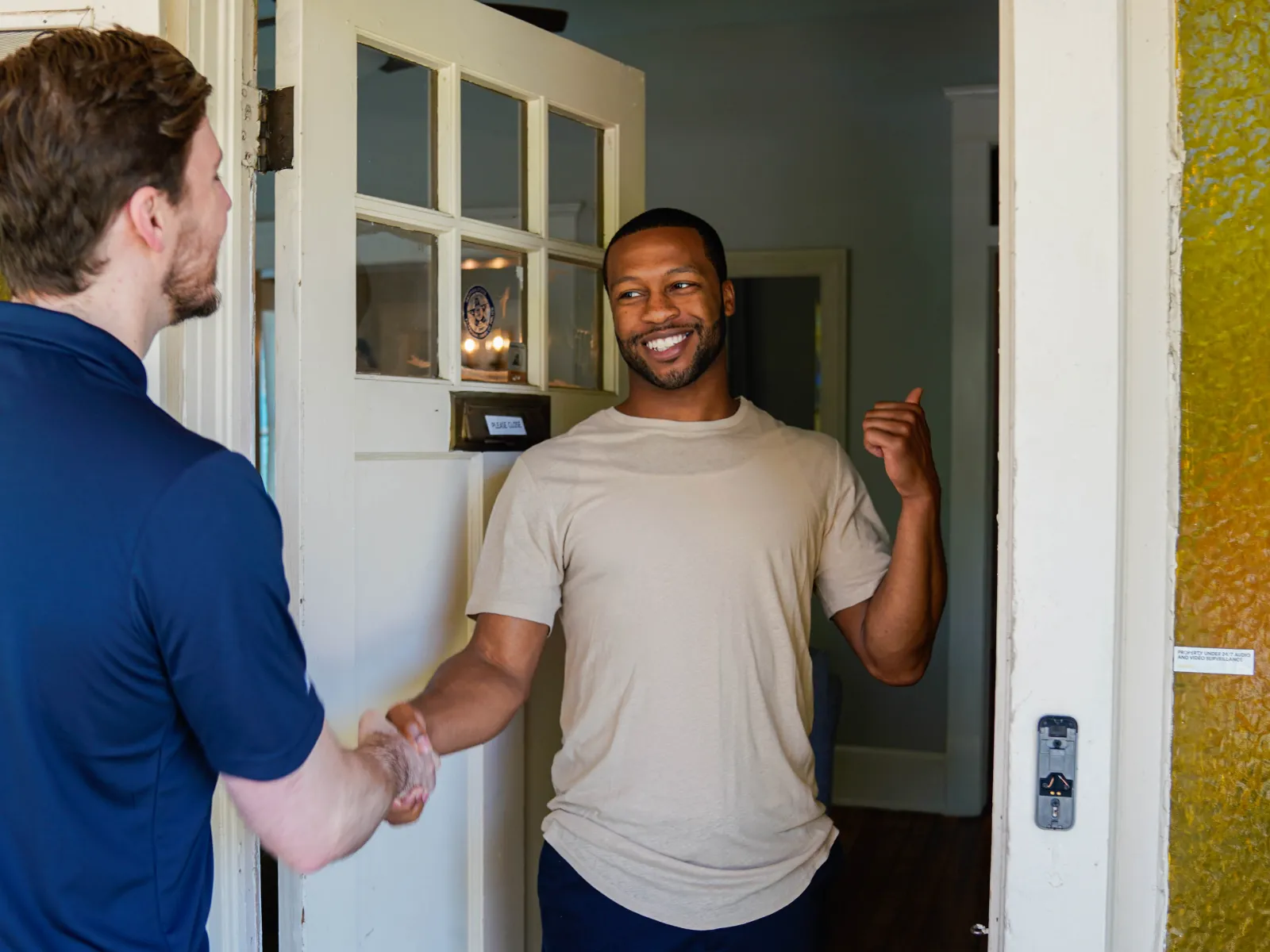 Two men shaking hands at the doorway, one smiling and welcoming the other inside a home.