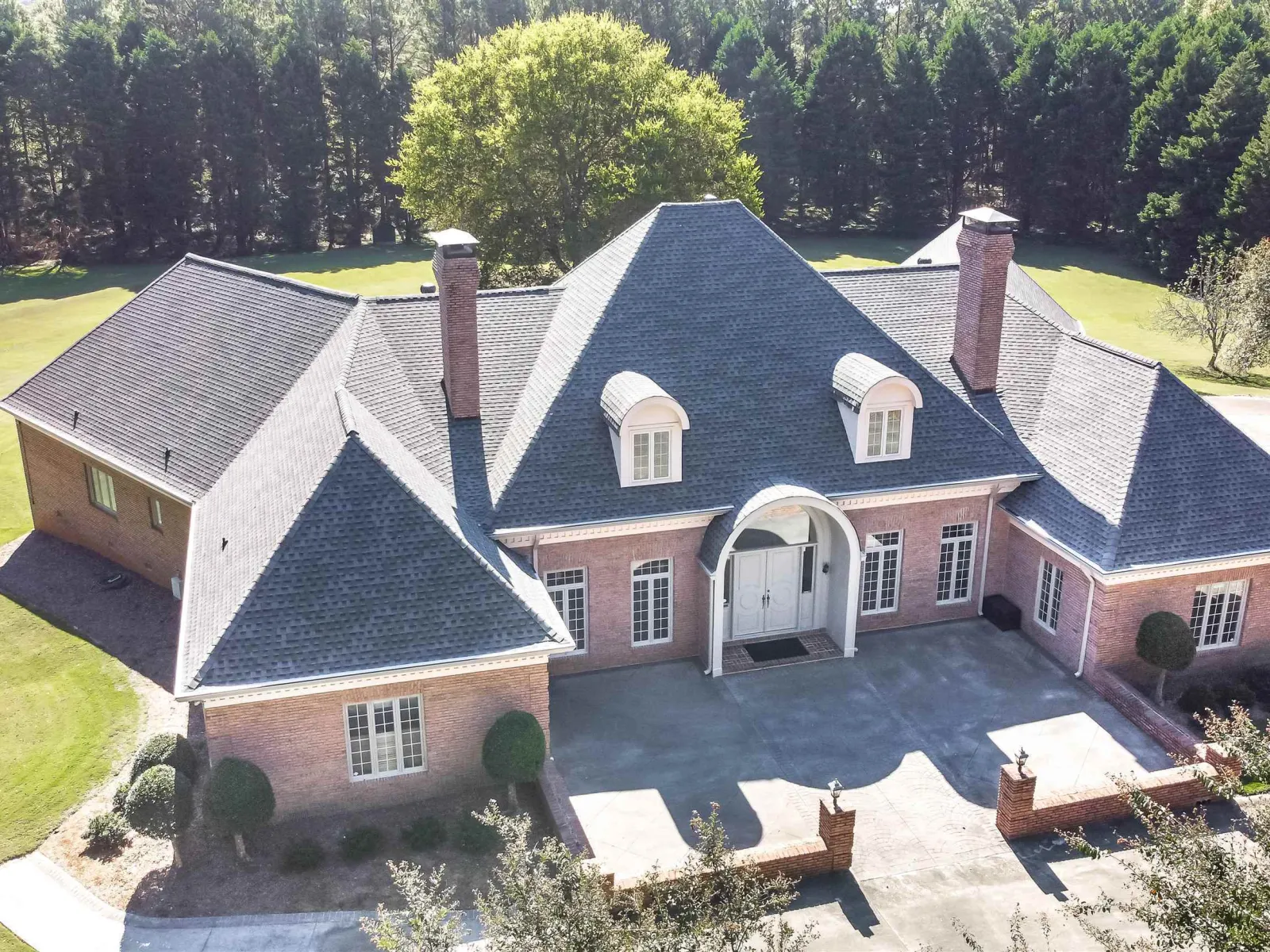 Large brick house with steep slate roof, three chimneys, arched front entrance, and manicured lawn surrounded by trees.