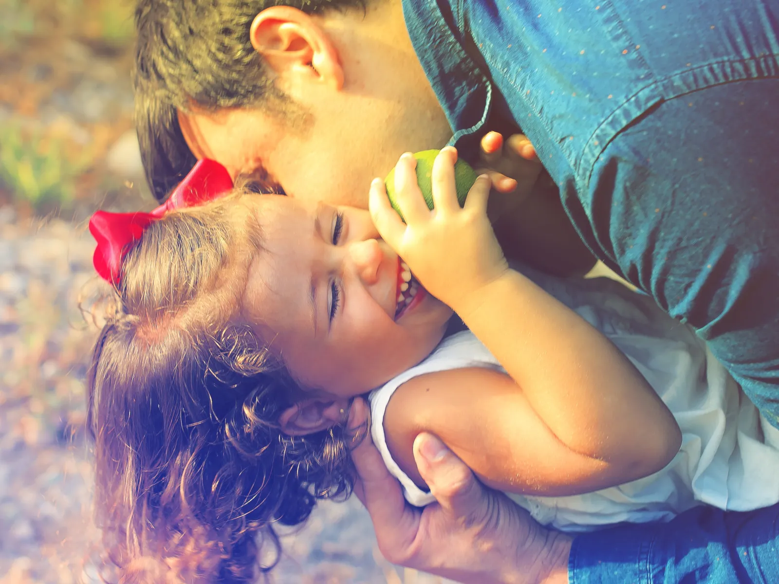 Father lovingly embraces and kisses smiling young daughter outdoors in warm sunlight.