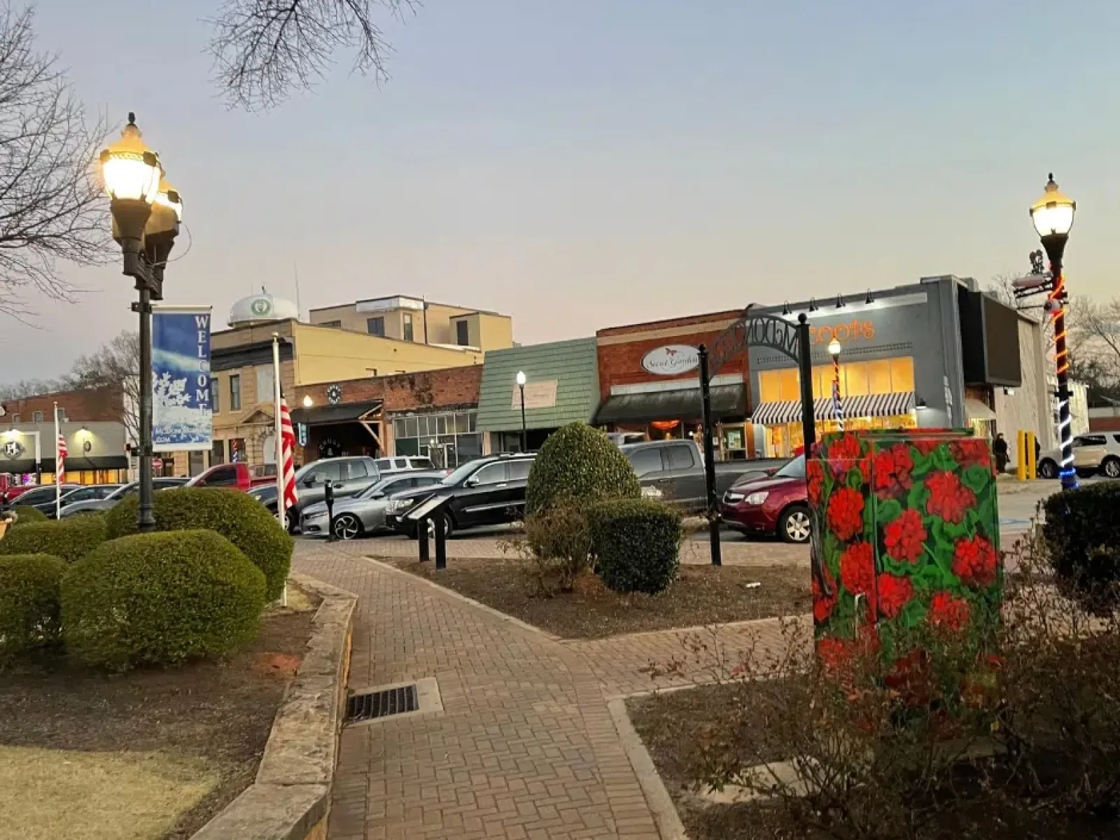 Small town street scene at dusk with shops, parked cars, trimmed bushes, and decorative lamp posts.