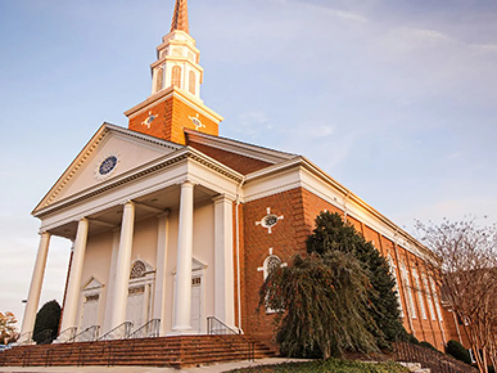 Red brick church with white columns and tall steeple under a clear blue sky on a sunny day