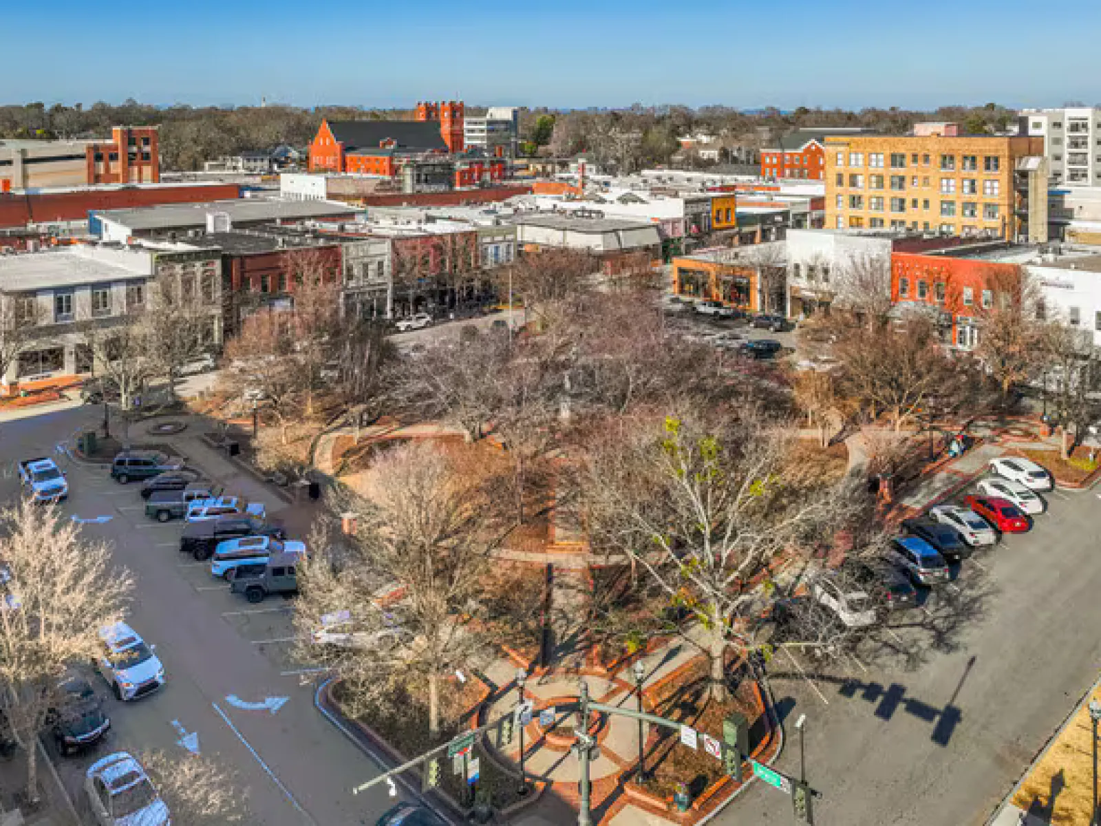 Downtown city square with leafless trees, parked cars, surrounding brick and historic buildings under clear sky