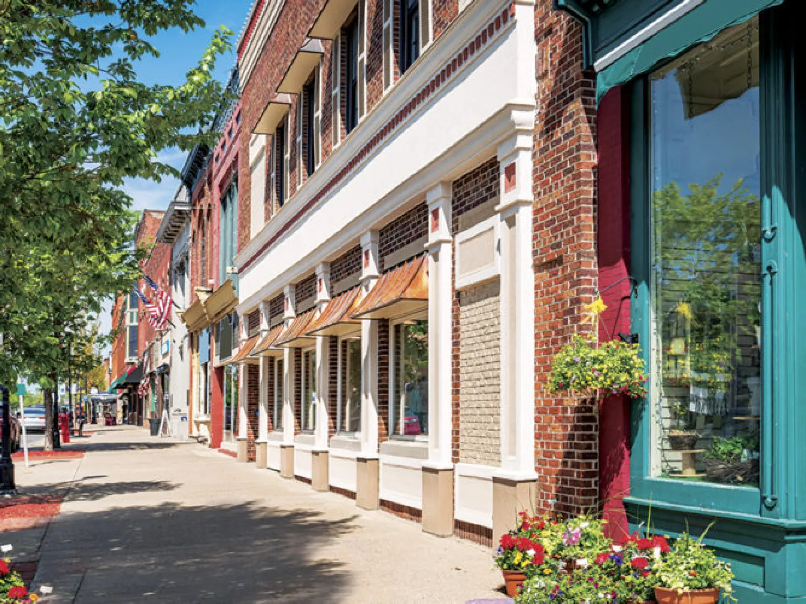 Sunny small-town street with brick buildings, shops, trees, cars, and colorful flower pots along the sidewalk