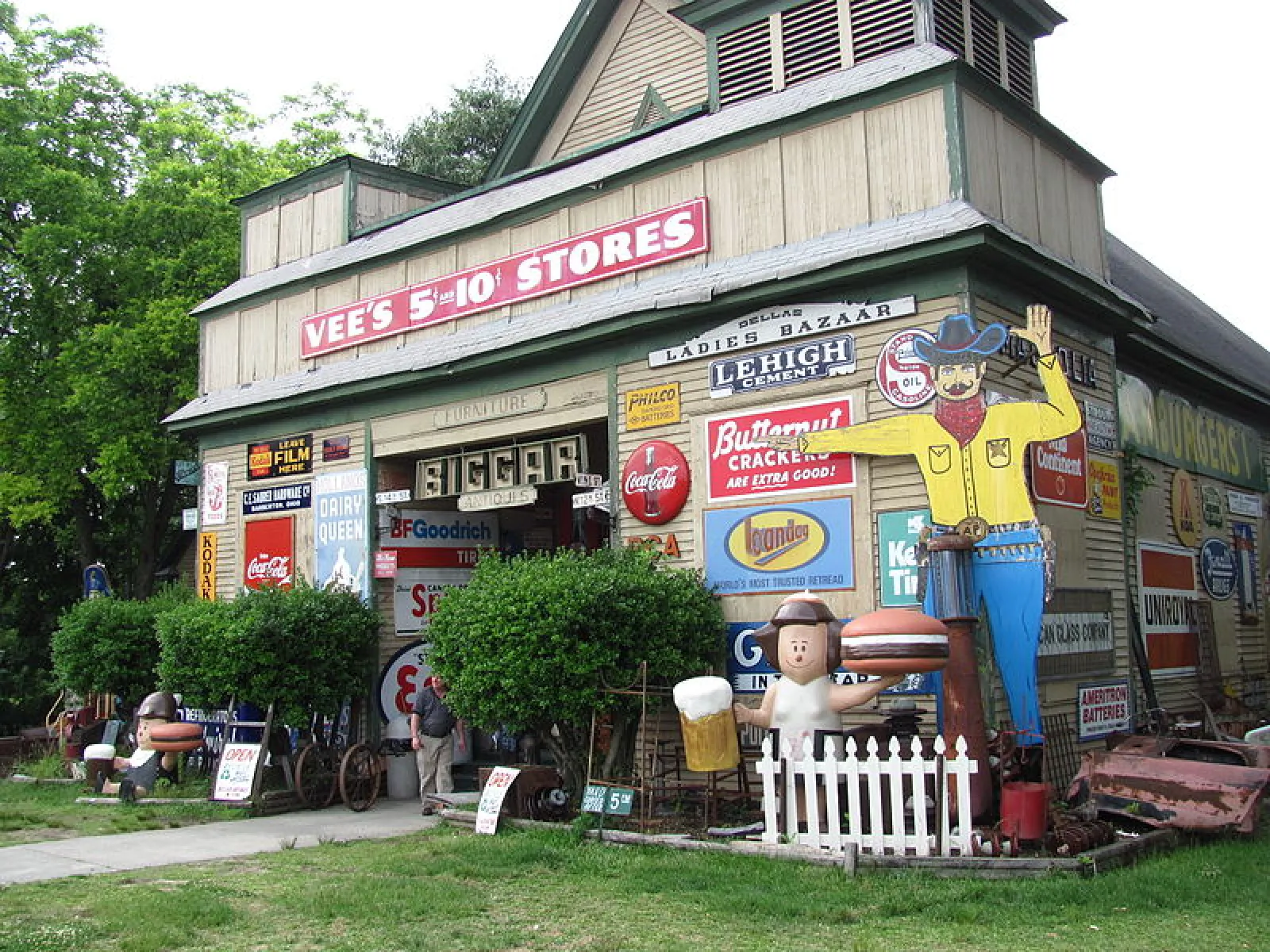 Old country store covered with vintage advertising signs and large wooden statues of a cowboy and woman with food.