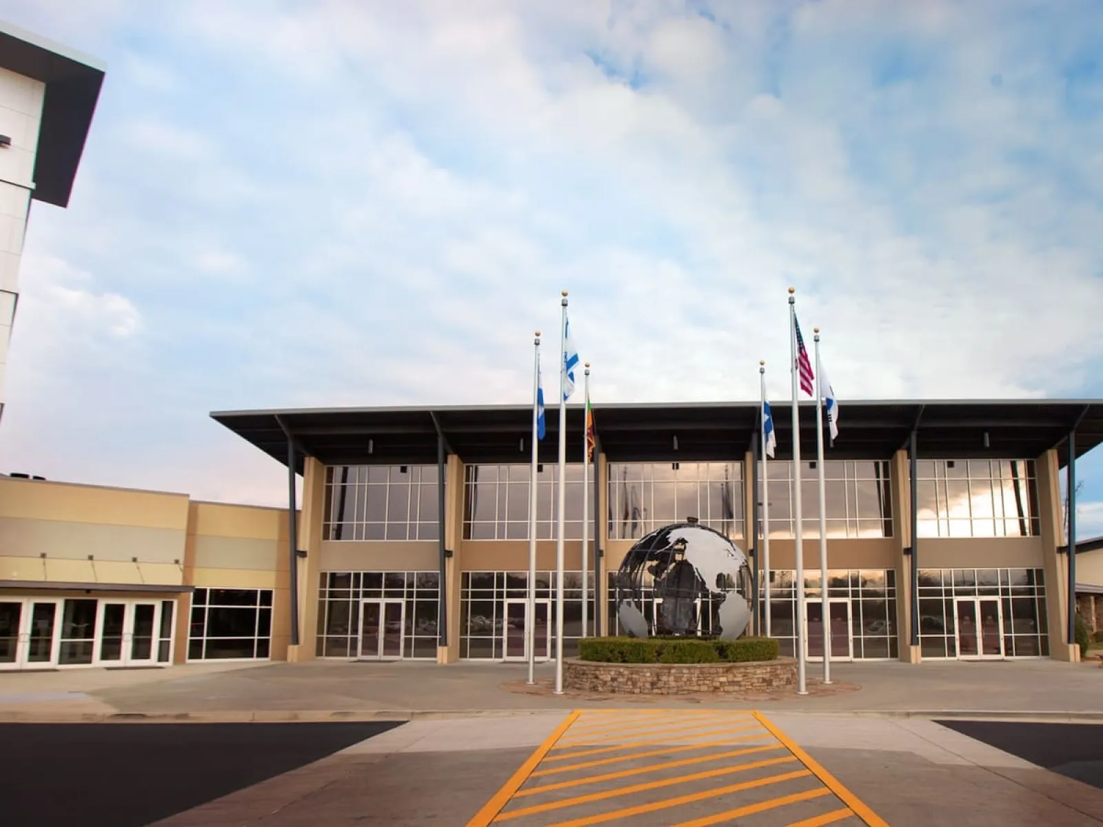 Modern church building with large glass windows, flagpoles, and a globe sculpture at the entrance under a cloudy sky.