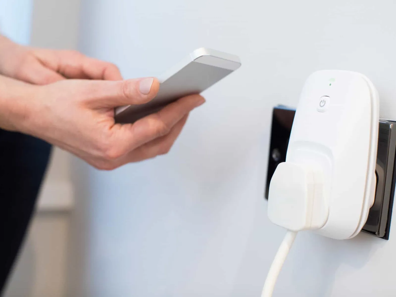 Hand holding smartphone near smart plug inserted in wall outlet for remote control of devices