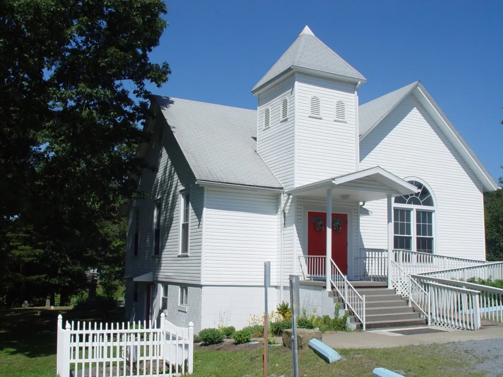 White church building with red double doors, a front porch, stairs, and a tower against clear blue sky.
