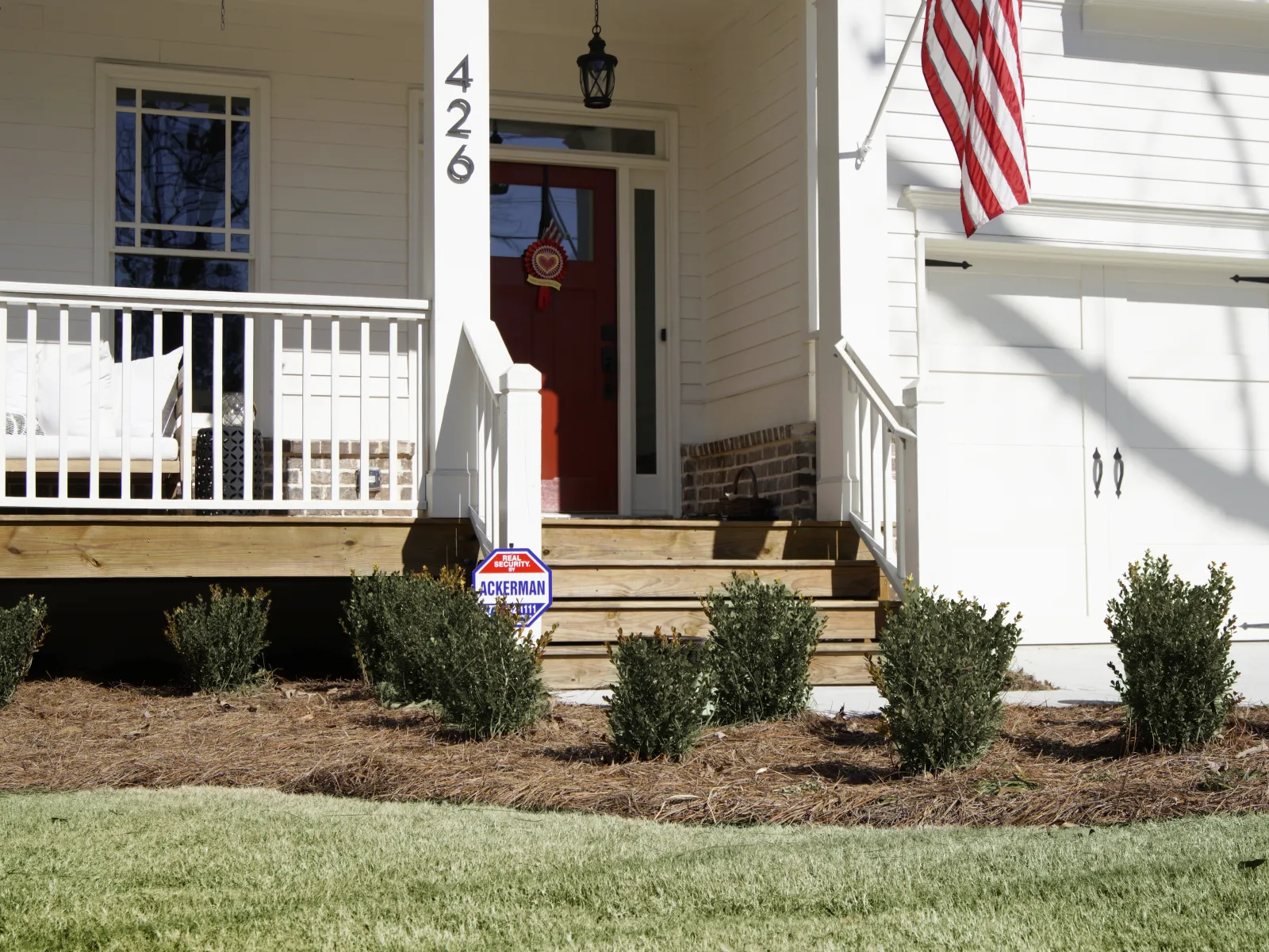 Front porch of a white house with red door, wooden steps, small bushes, and an American flag hanging by garage.