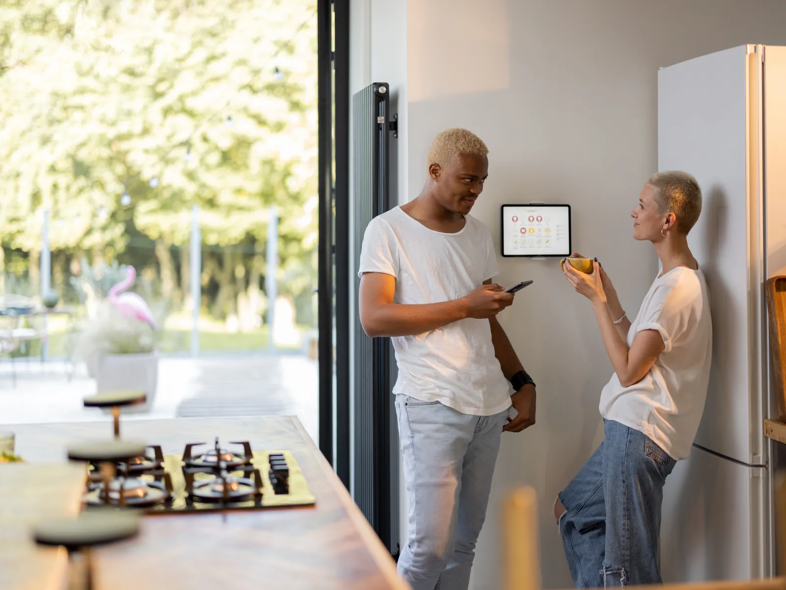Two people with light hair casually dressed standing in a kitchen by a wall-mounted smart home control panel.