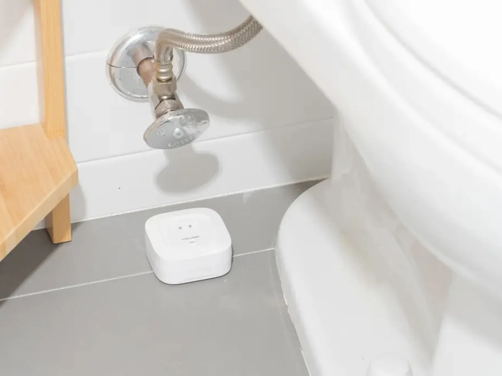 Small white water leak sensor installed on bathroom floor under toilet next to pipe and wooden shelf.