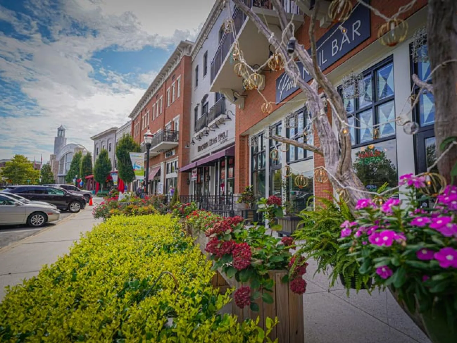 Colorful flowers line a vibrant downtown street with shops and parked cars under a partly cloudy sky.