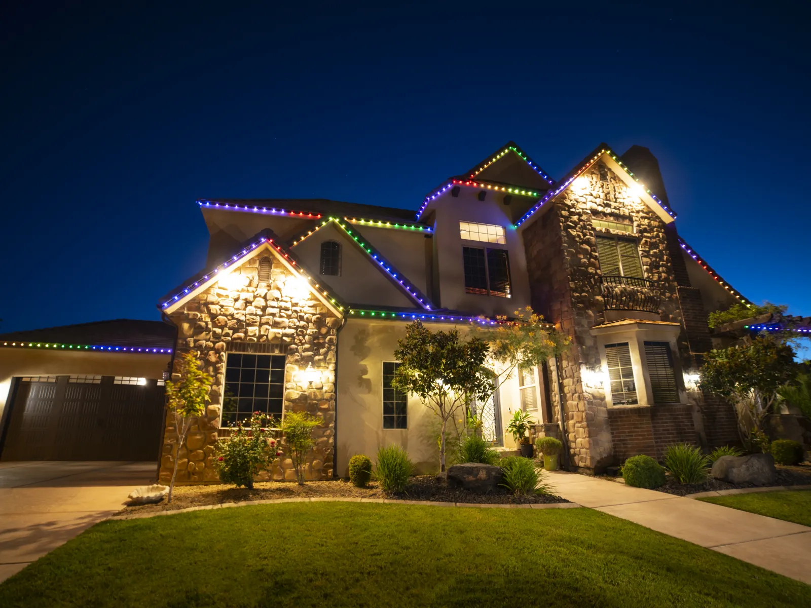 Large suburban house at dusk decorated with colorful Christmas lights on the roof and garden path.