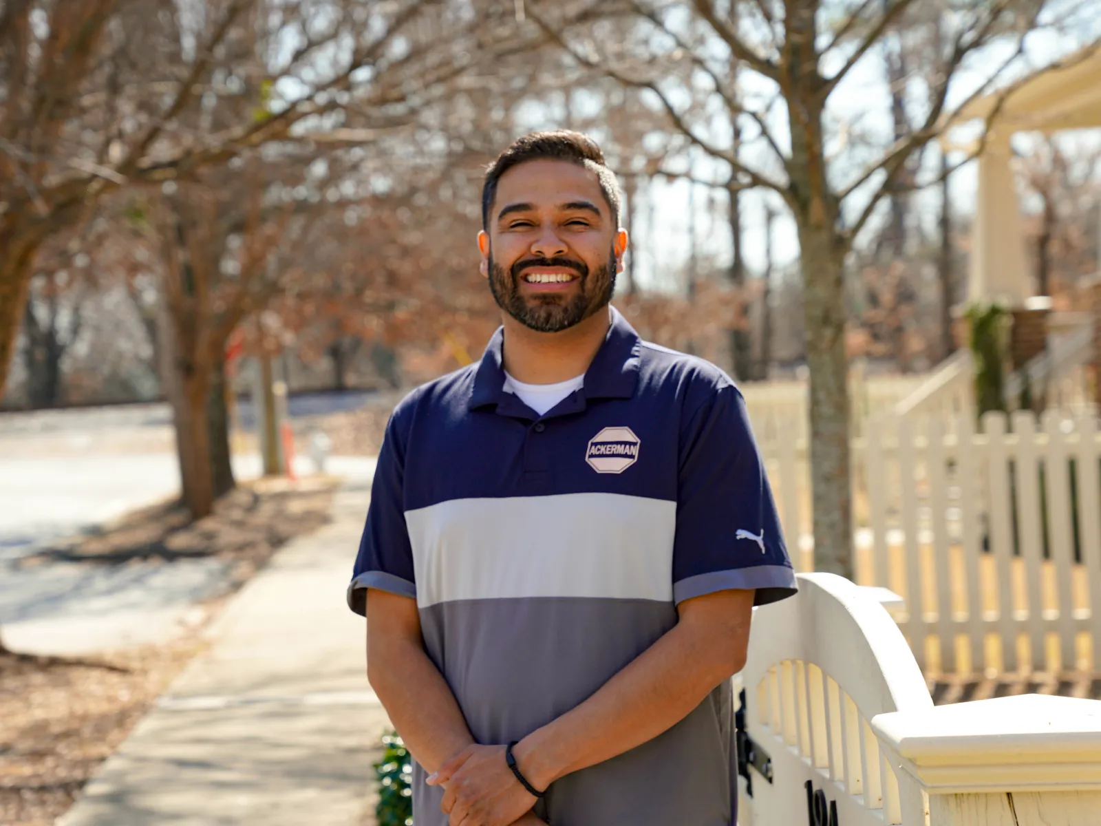 Smiling man in a blue and gray polo standing by a white picket fence on a sunny sidewalk with leafless trees.