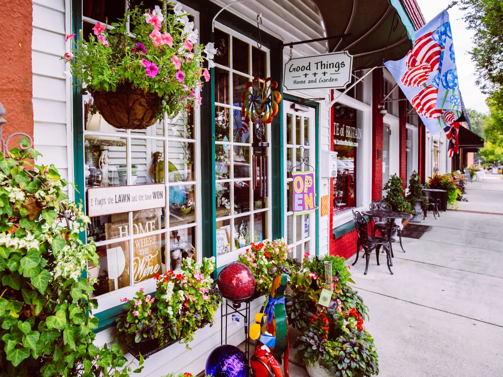 Charming storefront with hanging flower baskets, garden decorations, and American flags on a sunny sidewalk.