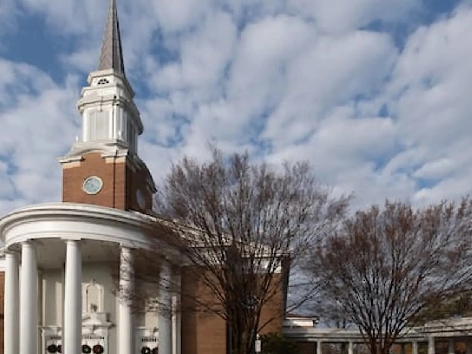 Traditional red brick church with white columns and a tall steeple under a cloudy sky.