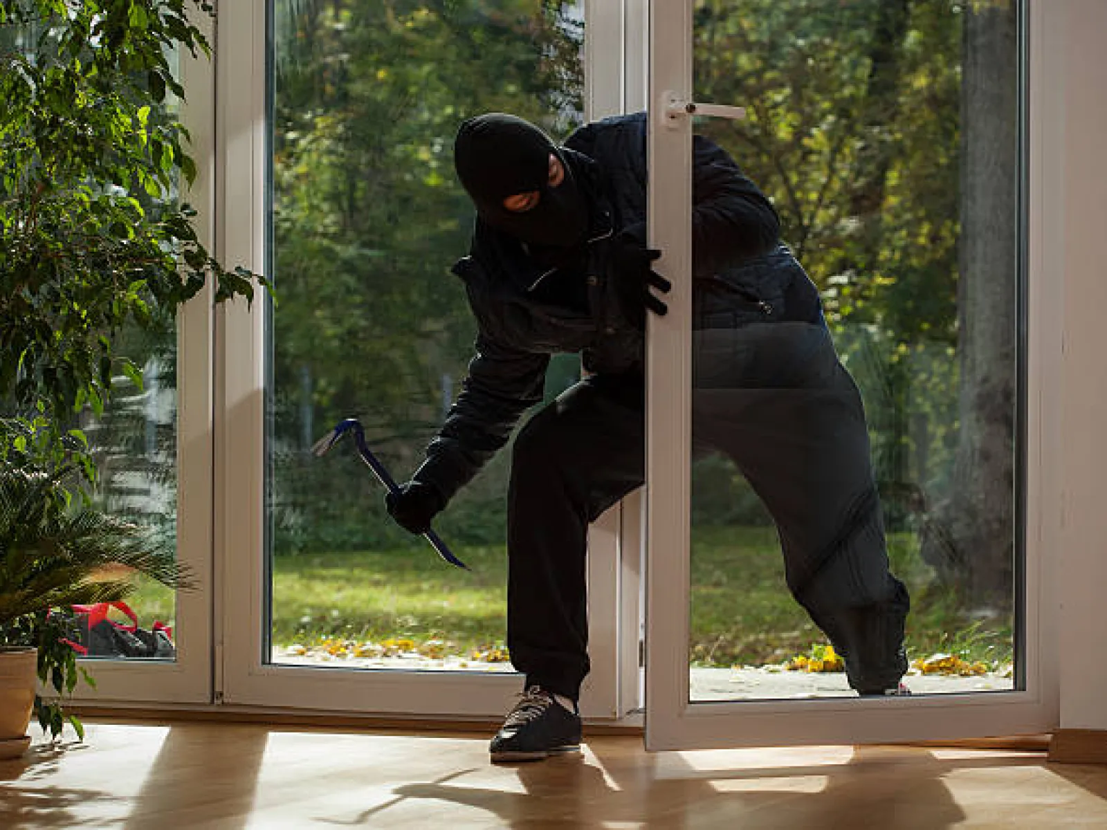 Masked intruder breaking into a house through a glass door holding a crowbar during daytime.