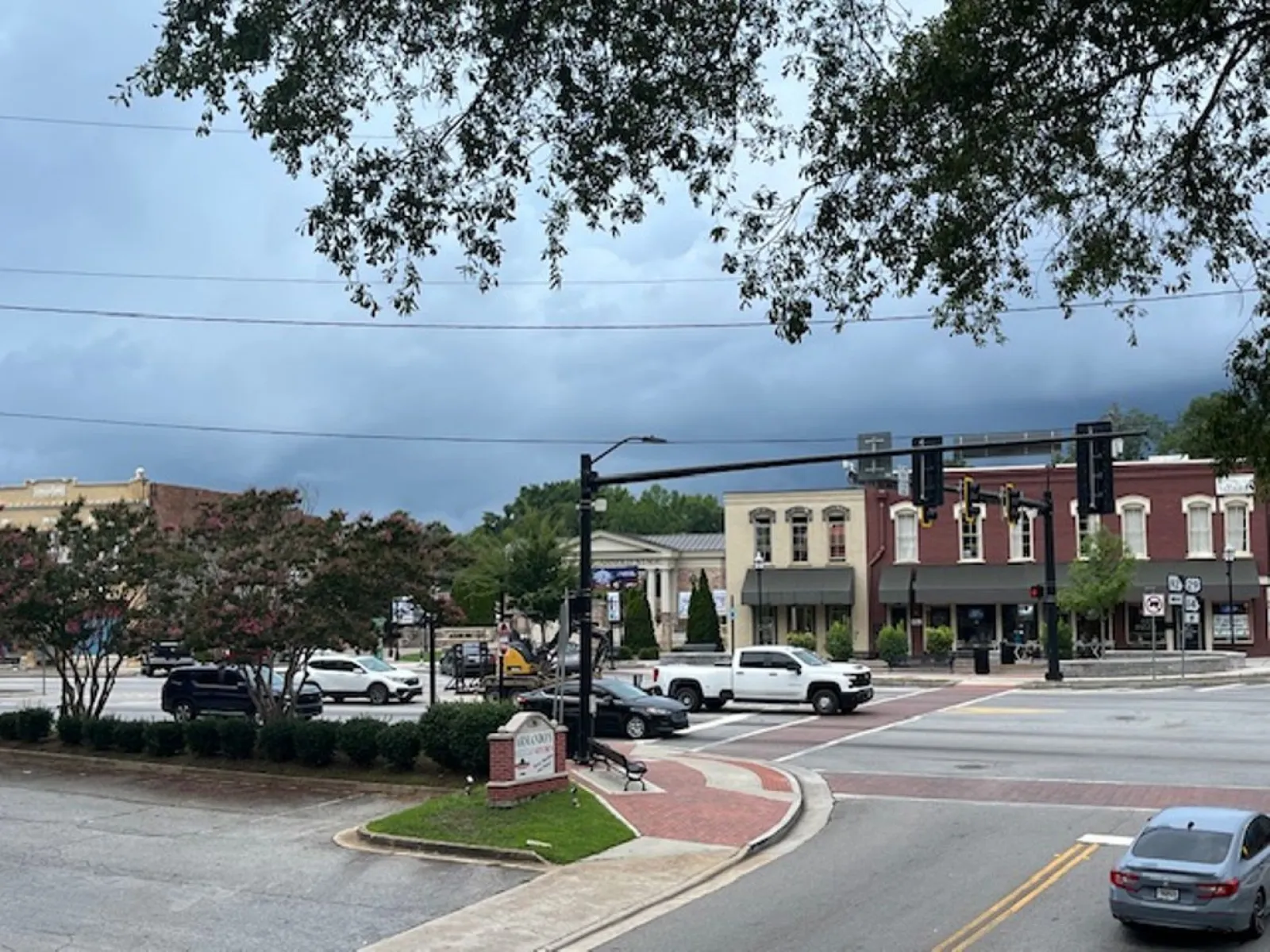 Small town street intersection with traffic lights, cars, old-style buildings, trees, and cloudy sky.