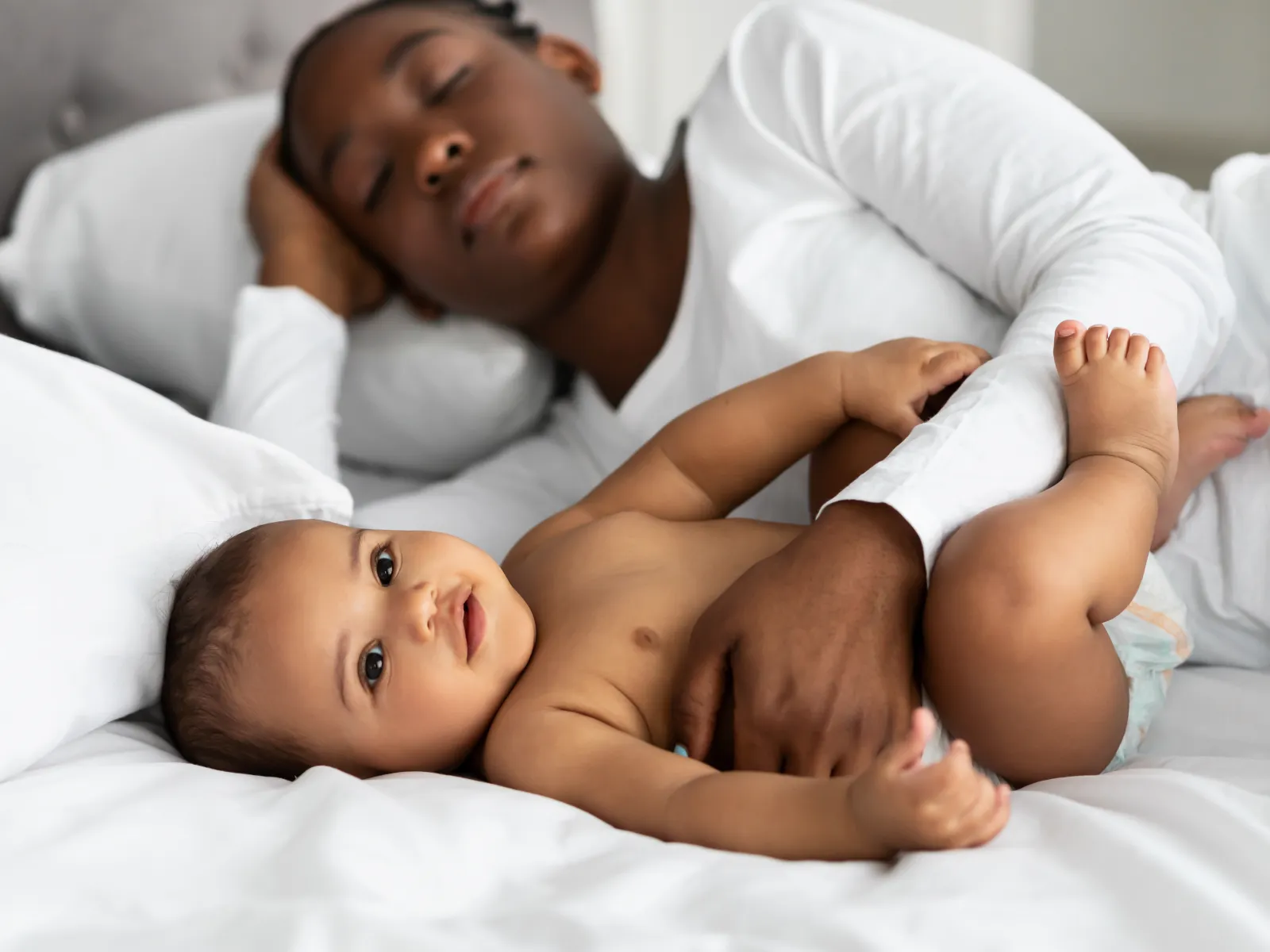 Mother asleep while holding baby lying on white bed sheets in a cozy bedroom setting.