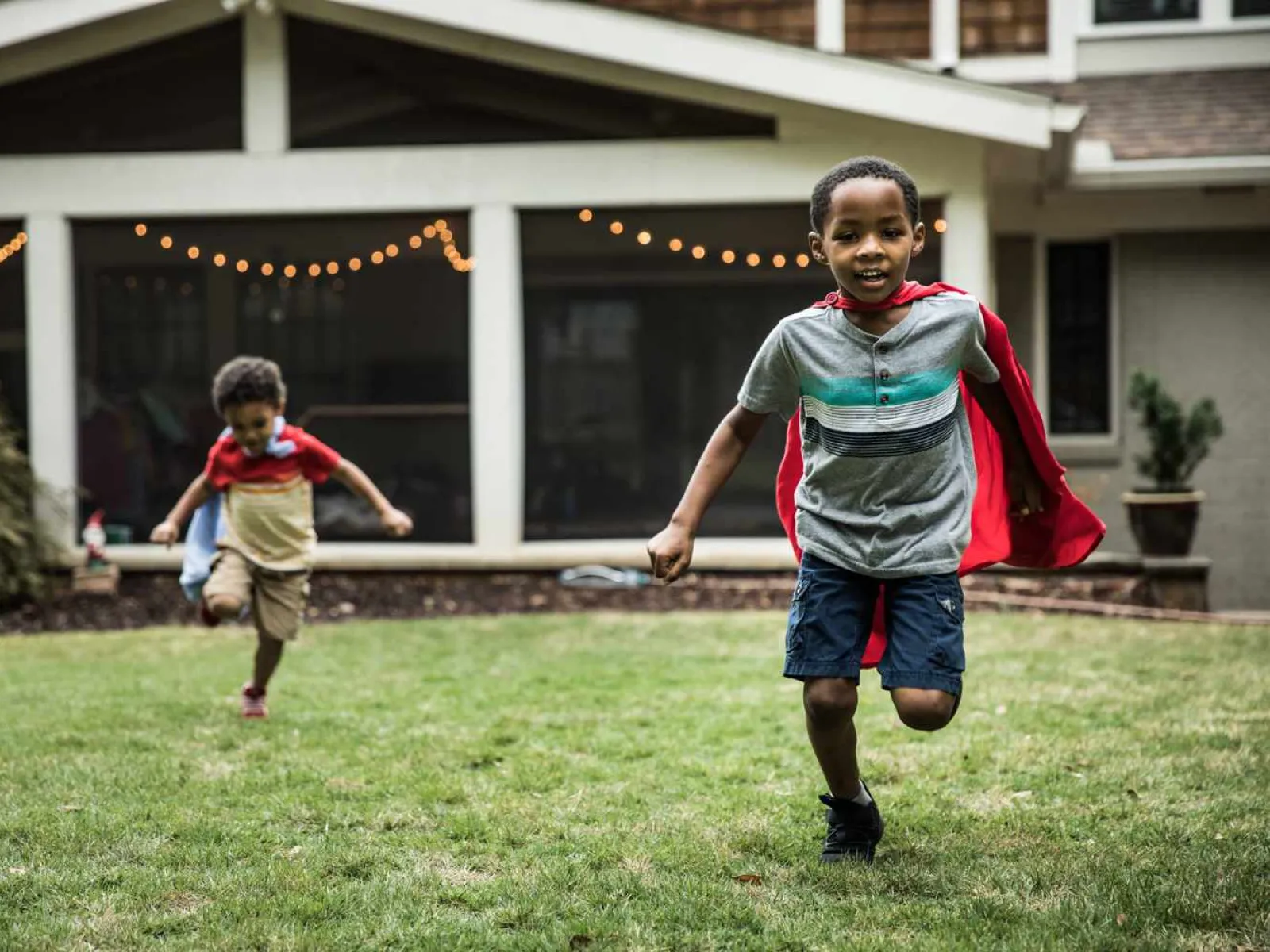 Two young boys wearing red capes running and playing on a grassy backyard with a house in the background