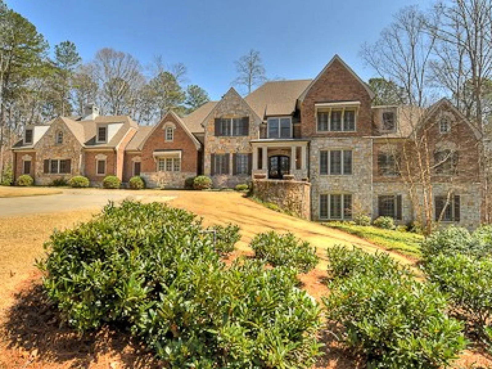 Large luxury stone and brick mansion with landscaped front yard and shrubs under clear blue sky.