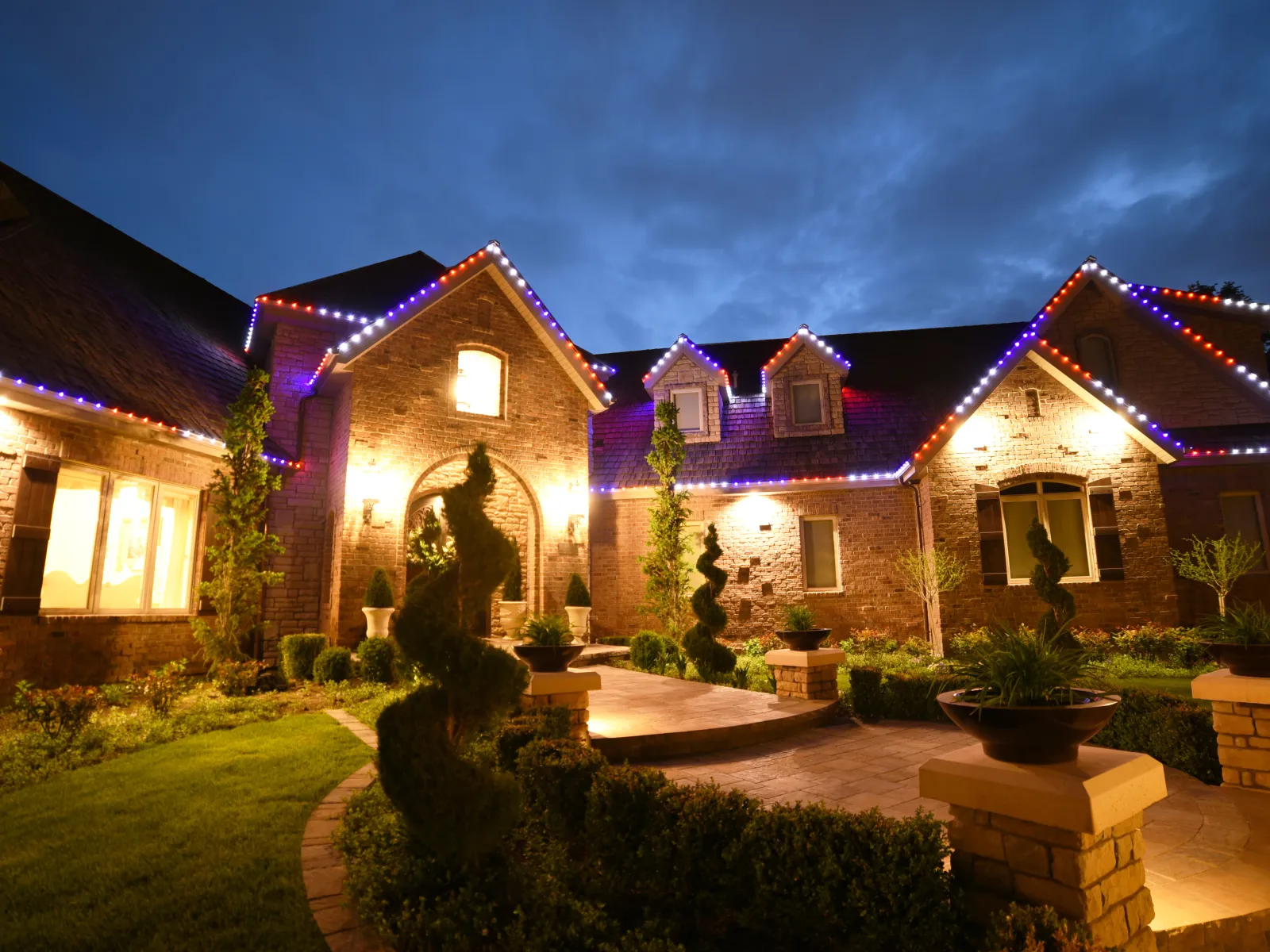 Large brick house illuminated with warm lights and colorful roofline LED decorations at dusk