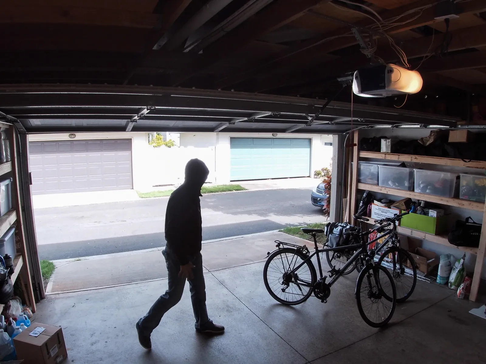 Person in hoodie walking inside a garage with bicycles and storage shelves near an open garage door.