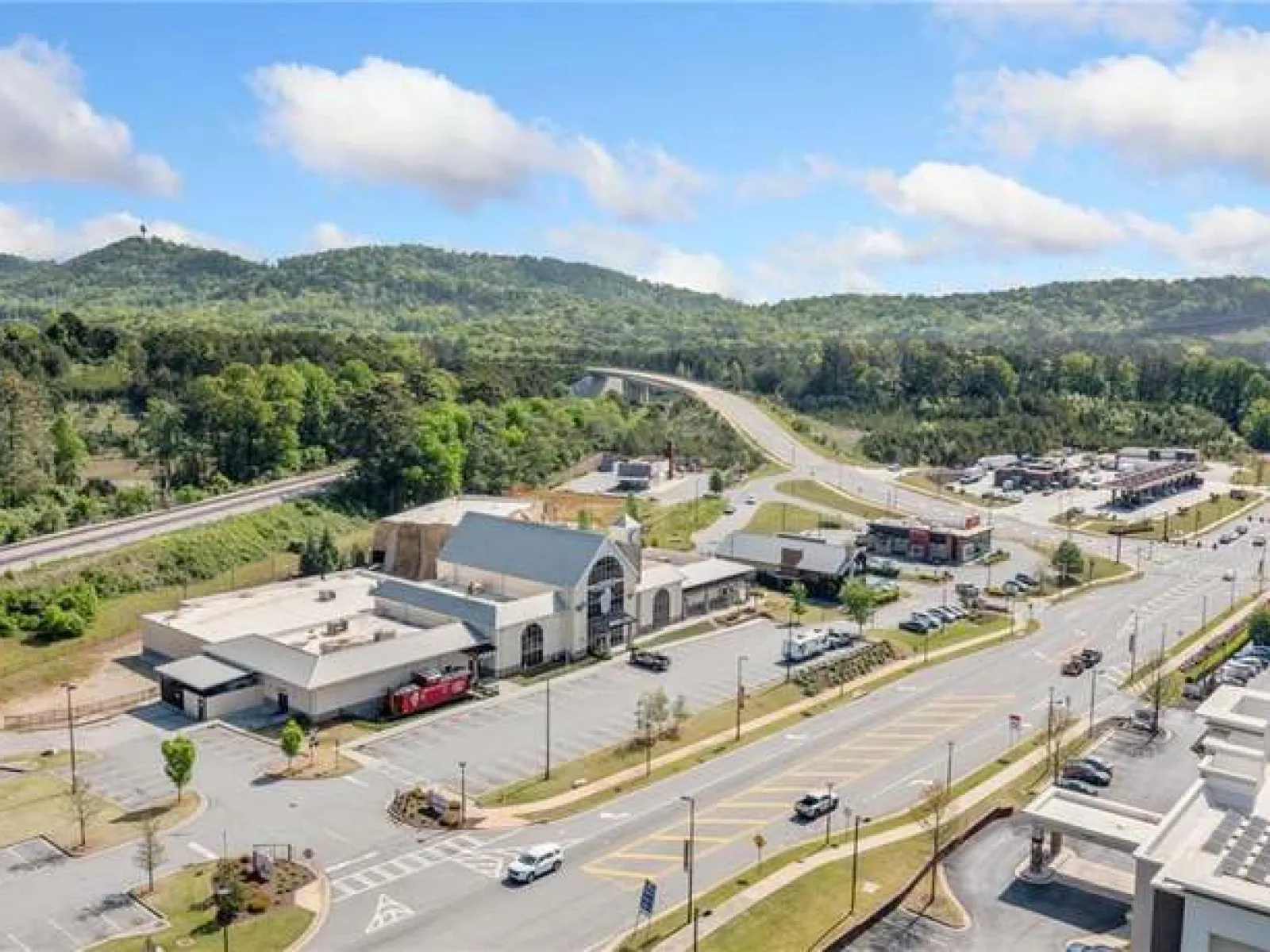 Aerial view of a commercial area with parking lots, roads, and green forested hills under a blue sky with clouds.