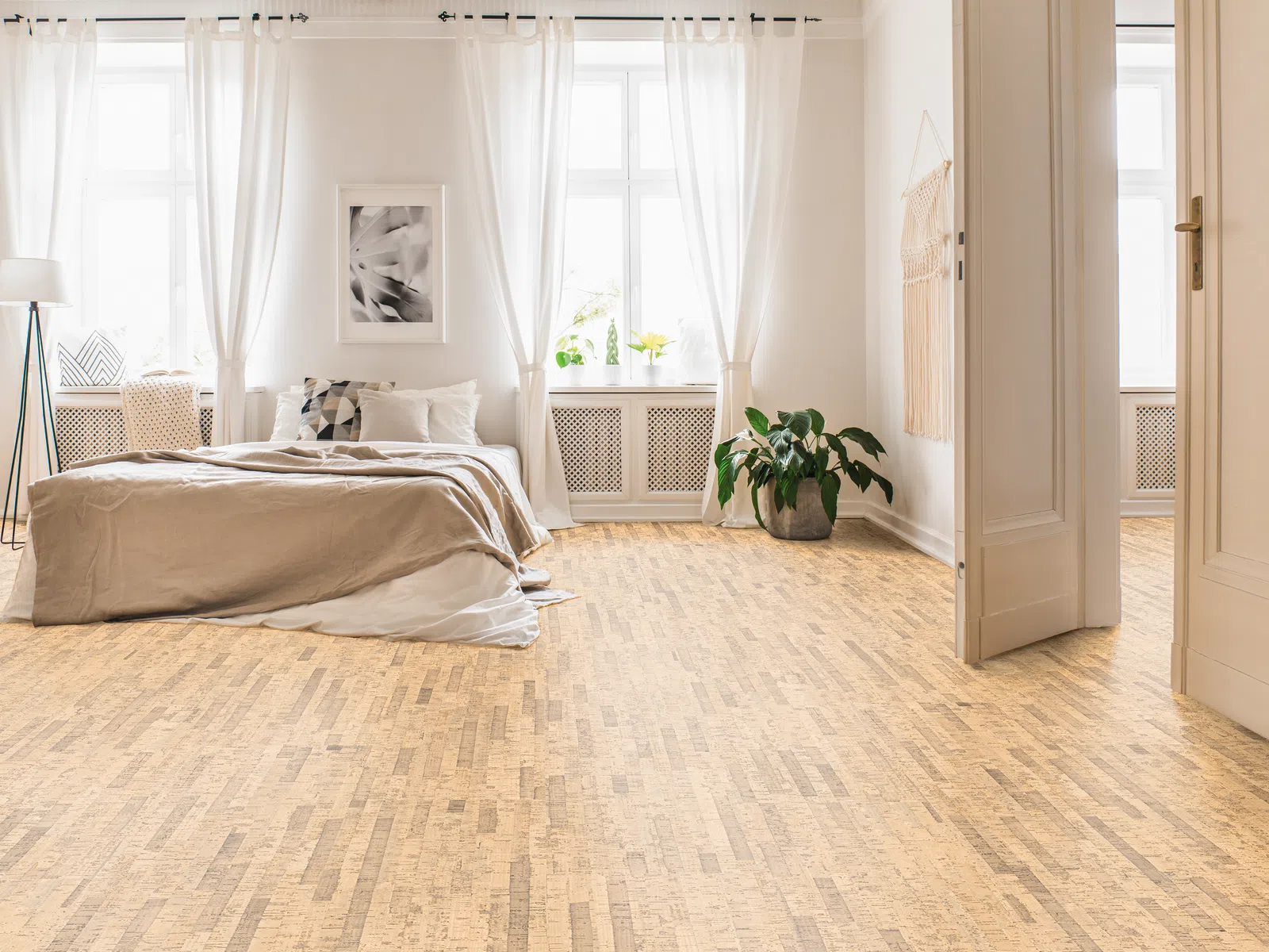 Bright bedroom with large windows, white curtains, beige bedding, wooden floor, and green potted plant