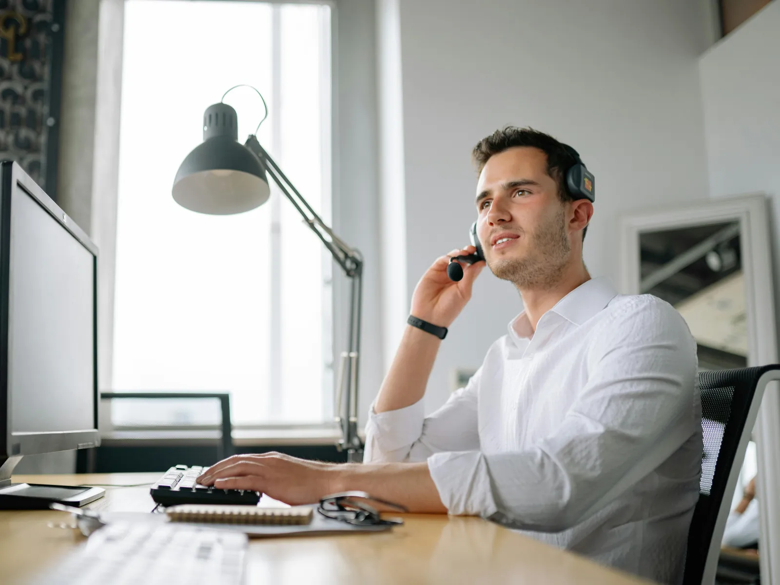 Man in white shirt using headset and computer at modern office desk with large window light.