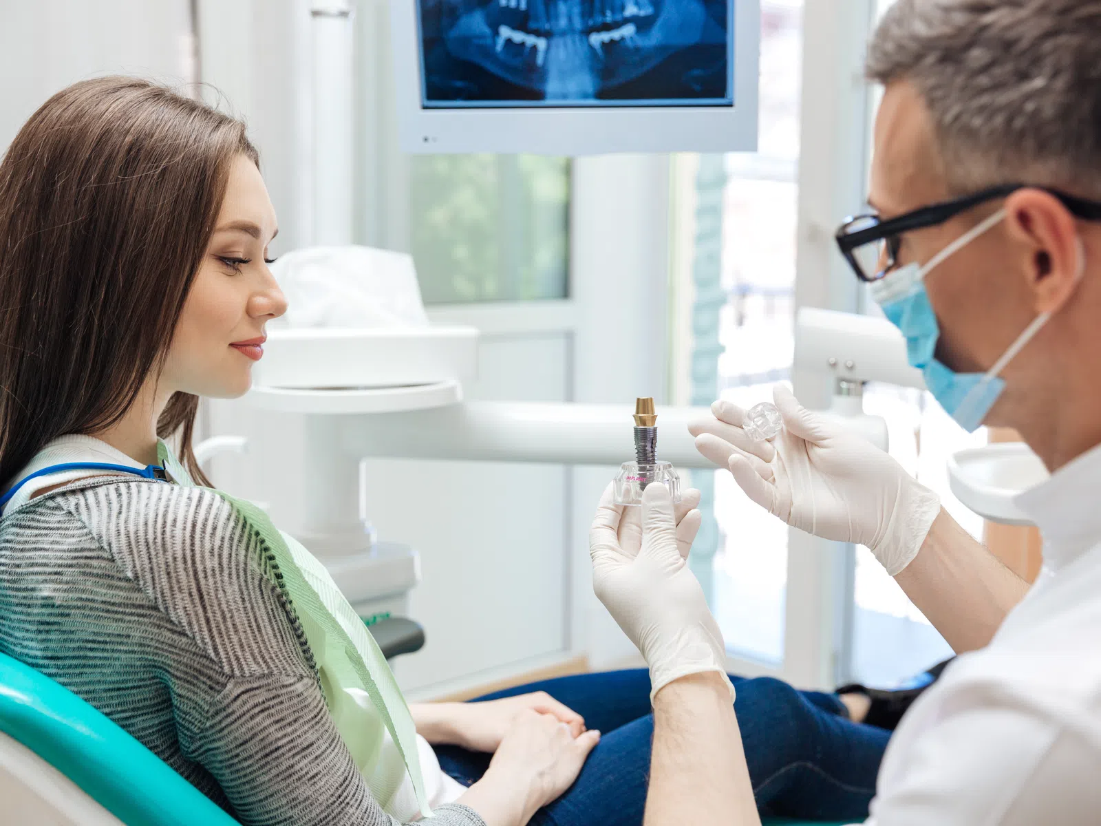 Dentist explaining dental implant procedure to female patient in dental office with X-ray visible.