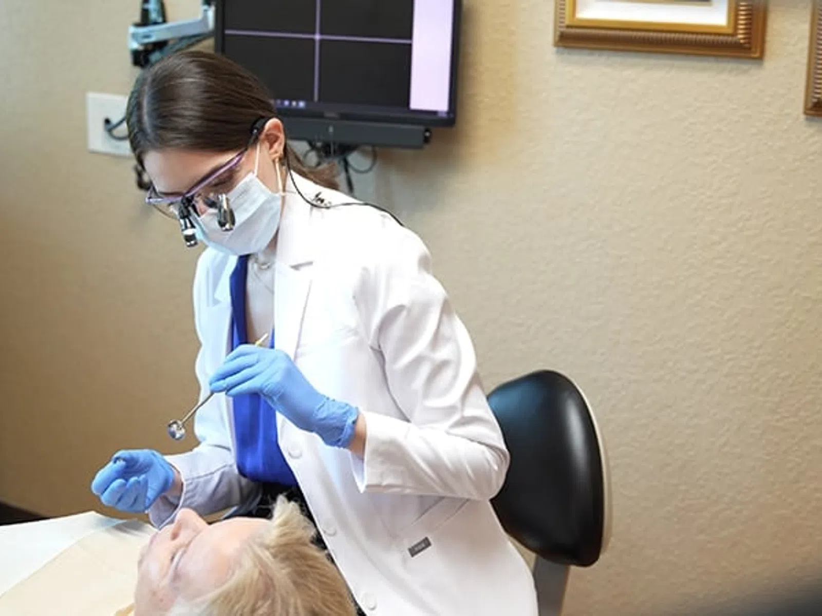 Female dentist in a white coat and mask examining an elderly patient's teeth in a dental clinic.
