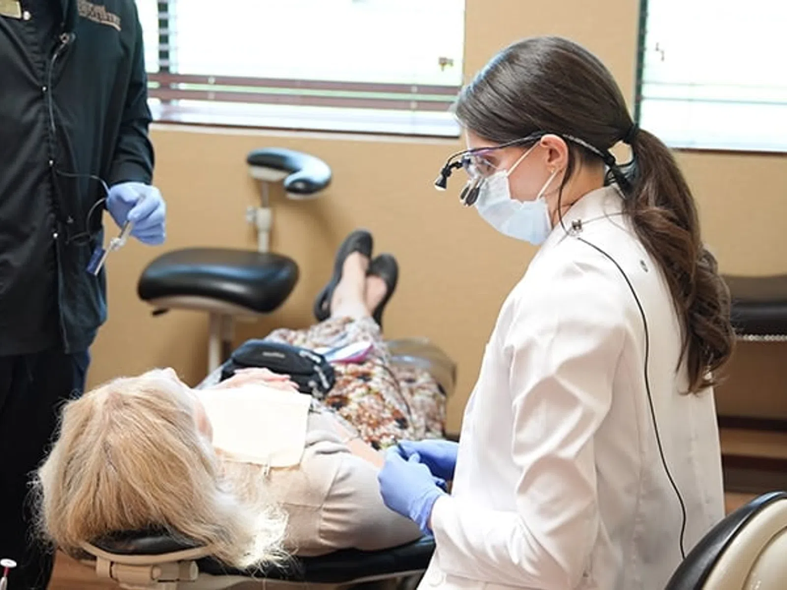 Dentist wearing a mask and gloves examining an elderly female patient lying in a dental chair in a clinic.