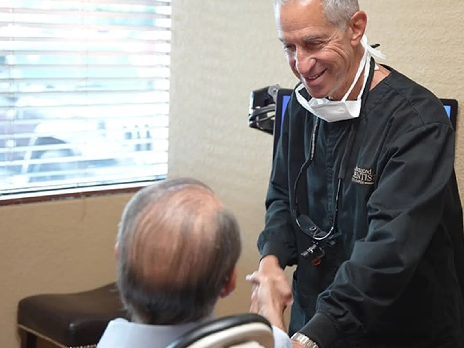 Dentist warmly greeting a senior male patient in a modern dental office with window blinds in the background